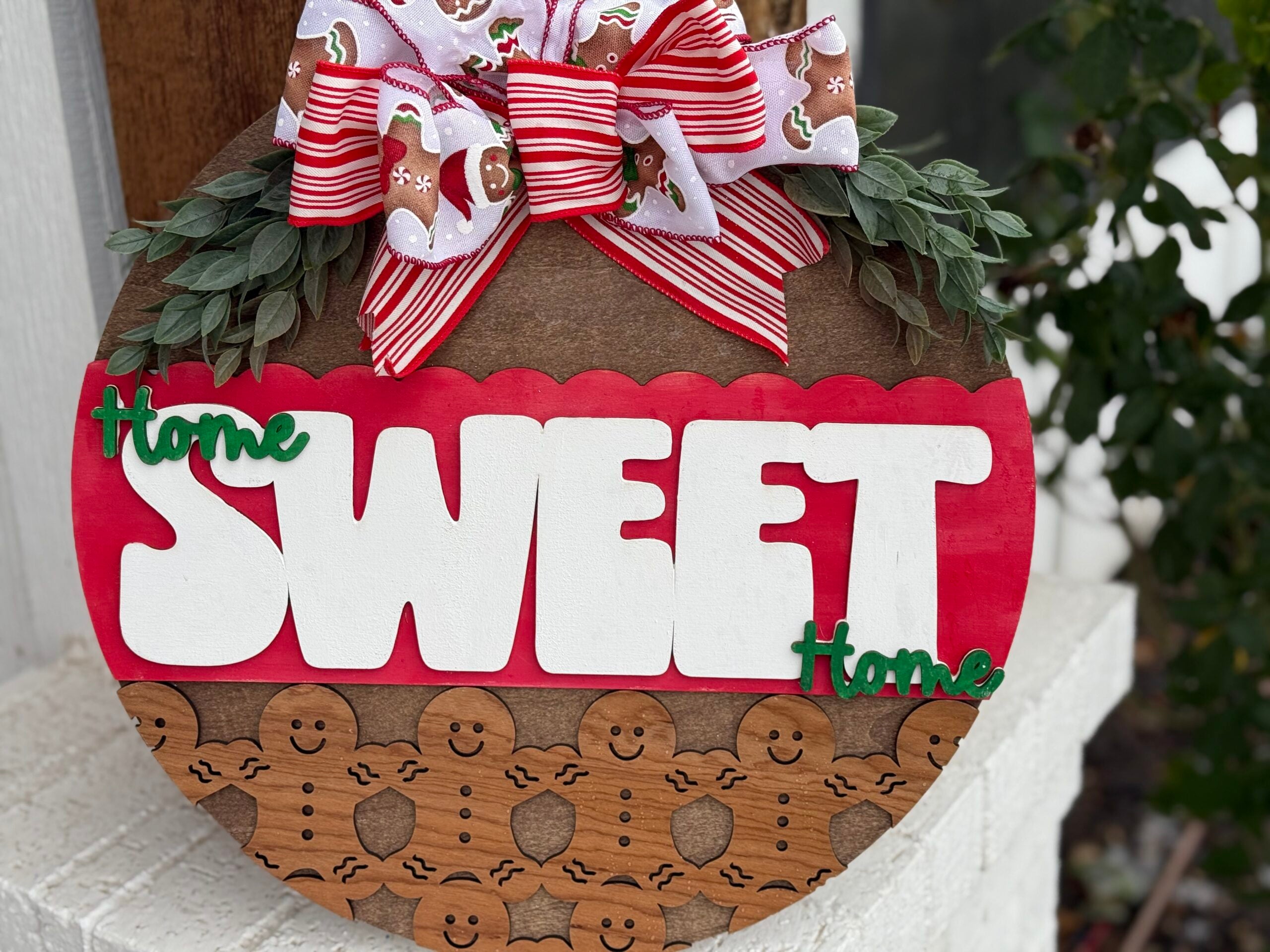 A round wooden sign adorned with green leaves and a large red, white, and gingerbread-patterned bow at the top. The sign reads Home Sweet Home in white and green letters, with a gingerbread men pattern on a brown section below the text.