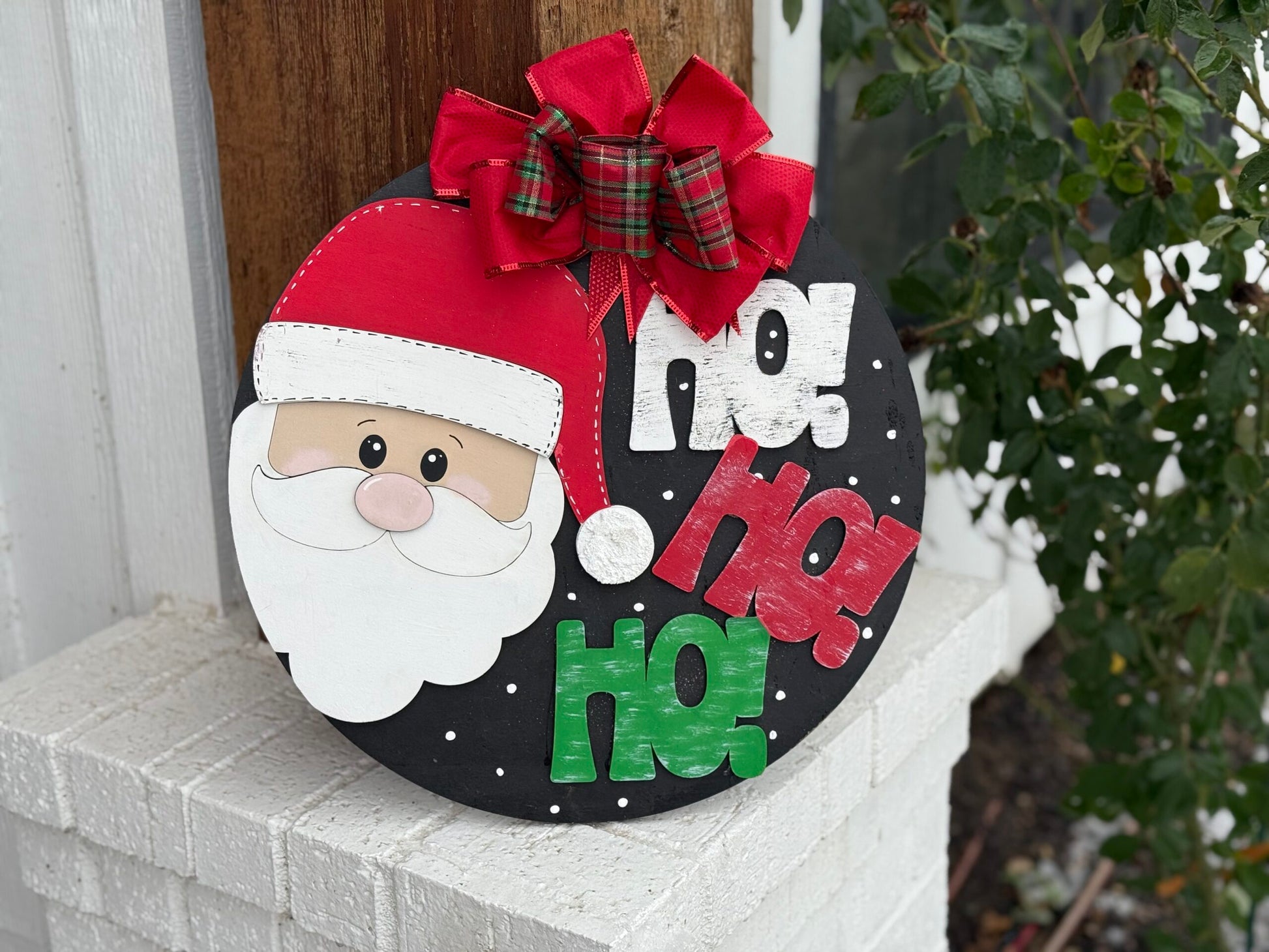 A festive circular sign features Santas face with a red hat and a fluffy white beard. The background is black with white dots, and the words Ho! Ho! Ho! in white, red, and green text. A plaid red and green bow sits at the top. The sign leans against a white brick column.