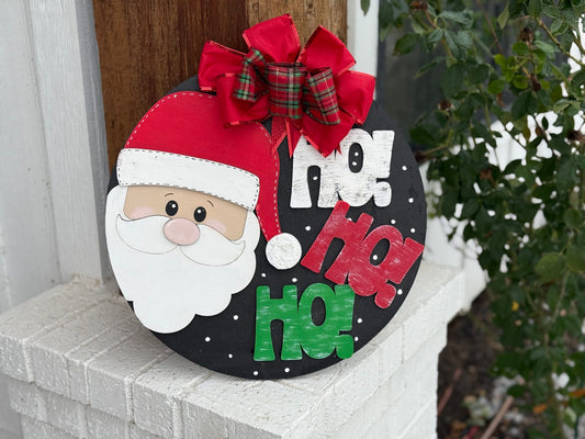 A festive circular sign features Santas face with a red hat and a fluffy white beard. The background is black with white dots, and the words Ho! Ho! Ho! in white, red, and green text. A plaid red and green bow sits at the top. The sign leans against a white brick column.