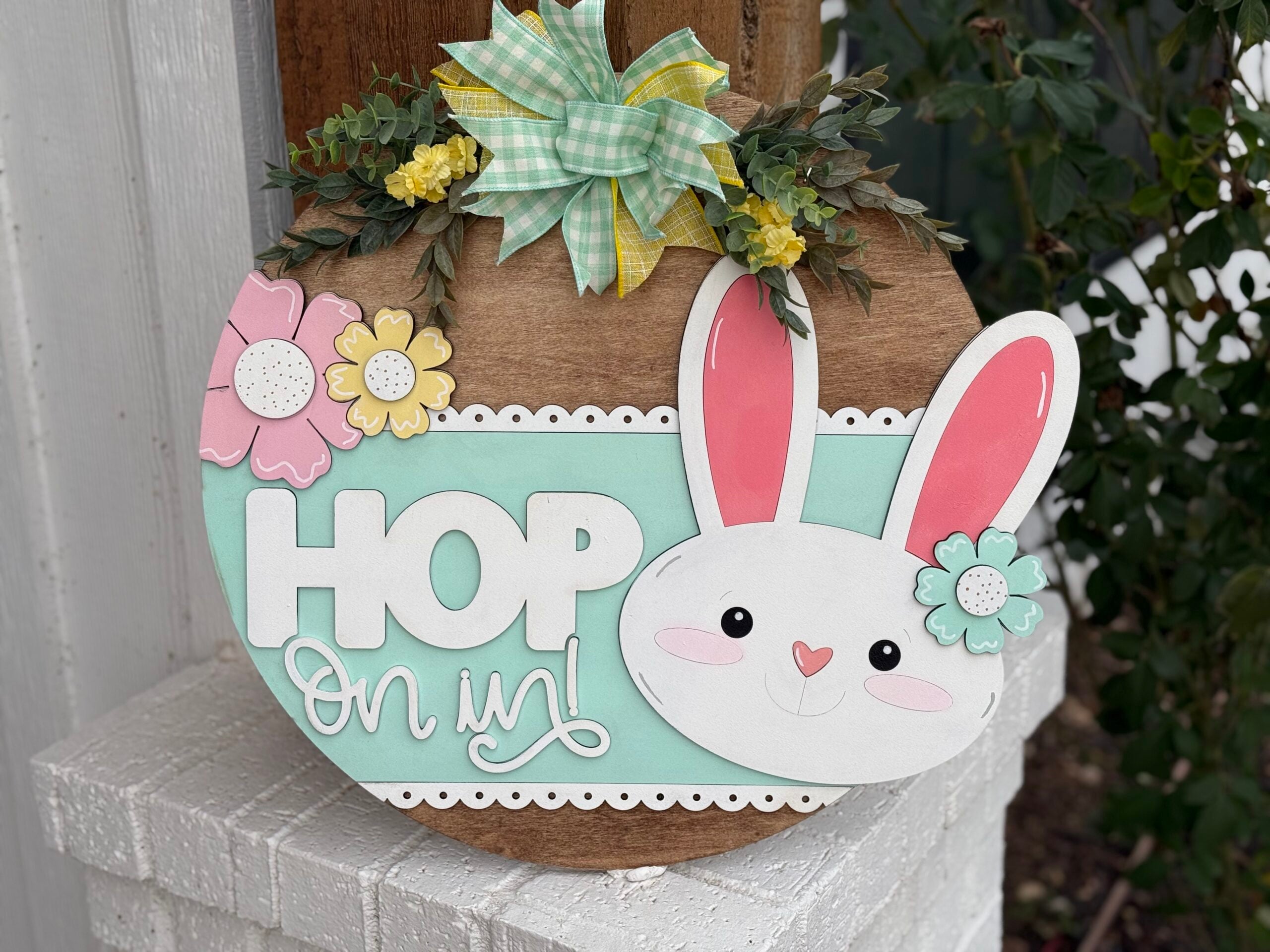A decorative Easter sign shaped like an egg features a cute white rabbit face with pink ears, surrounded by pastel flowers, green leaves, and a plaid bow. The words HOP On in! are displayed in white, with a backdrop of light blue and wood. Its hanging on a white brick wall.