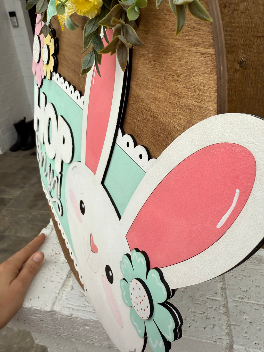 Close-up of a decorative wooden sign featuring a cartoon bunny with large pink ears and rosy cheeks. The bunny is outlined with pastel flowers and greenery, and the word HOP is cut out in bold letters. A hand is partially visible, holding the side of the sign.