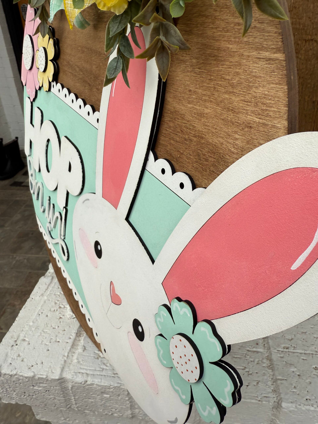 A close-up of a decorative wooden sign featuring a cute cartoon bunny with long pink ears and a cheerful smile. The bunny is adorned with colorful flowers. The word HOP is partially visible behind the bunny. Green leaves and pastel colors add a springtime feel.
