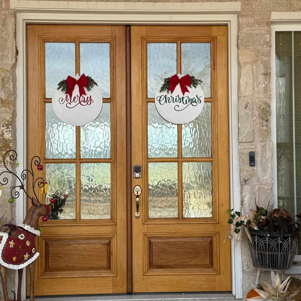A set of wooden double doors decorated for Christmas. Each door has a round white wreath with a red bow; the left wreath says Merry, and the right one says Christmas. A plant and decorative stand sit to the right, with a reindeer statue to the left, in front of textured glass panels.