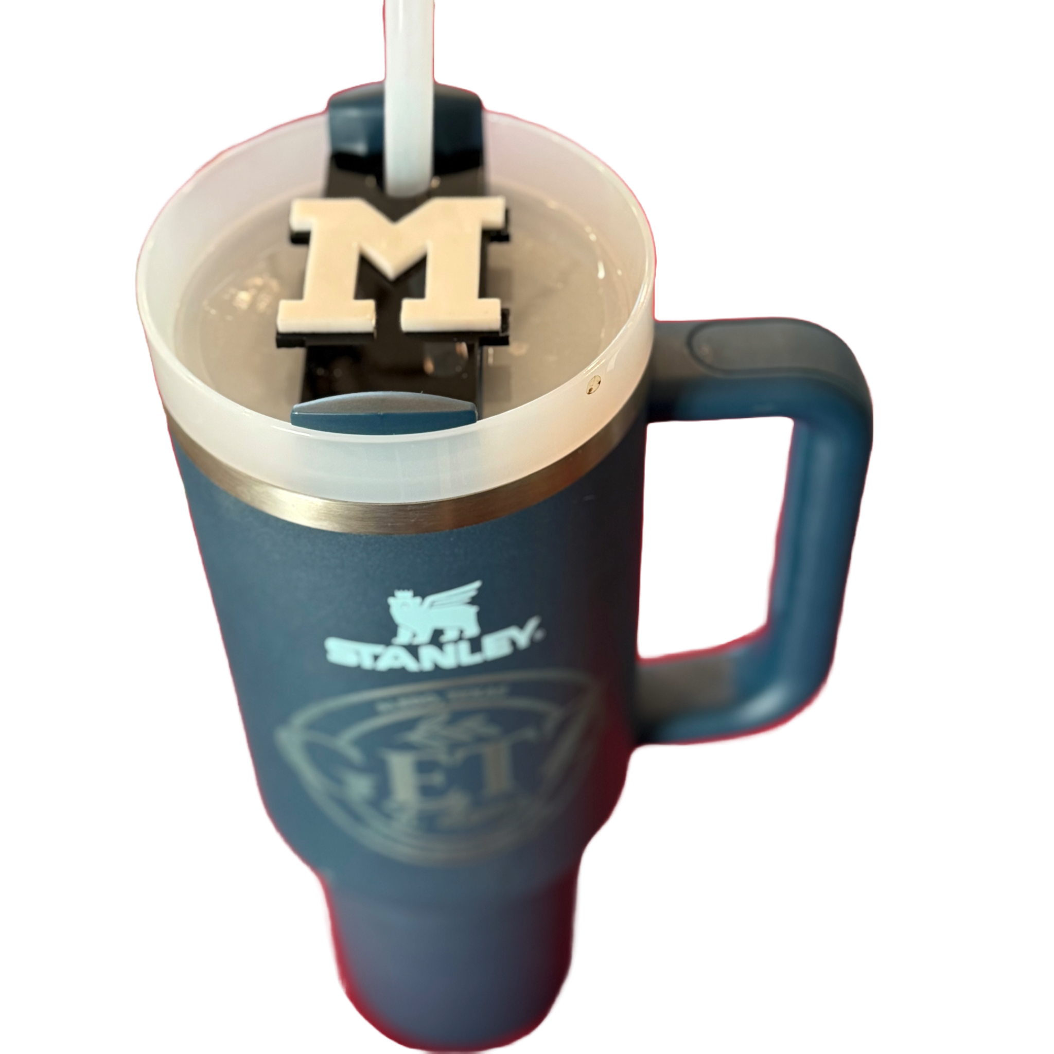 Blue Stanley cup with a handle, featuring a Mansfield High School logo on a black sliding lid. The lid has a straw inserted, and the cup contains a light-colored beverage. The Stanley logo is printed on the cup below the handle. The background is white.