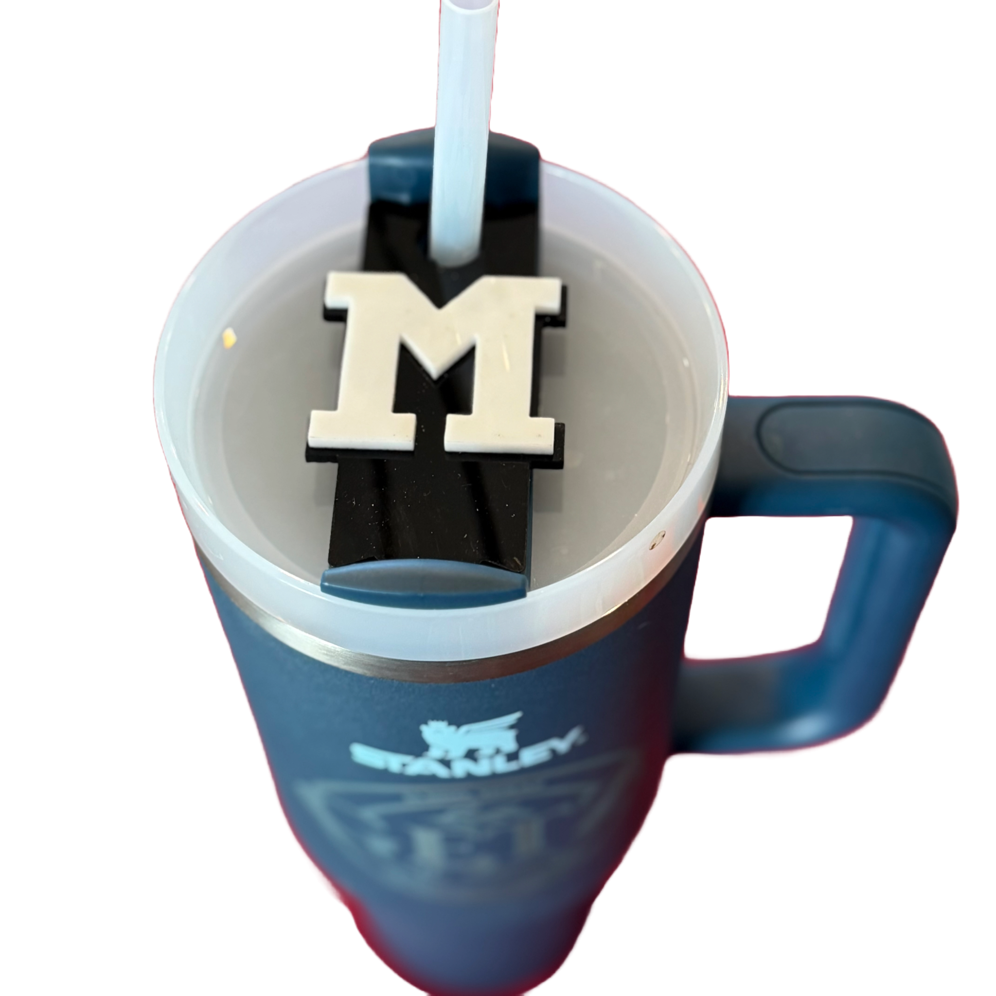 A blue Stanley tumbler with a handle features a white lid and a straw. On the lid, theres a black and white block letter M embellishment. The Stanley logo is visible on the tumblers side. The image is taken from above against a white background.