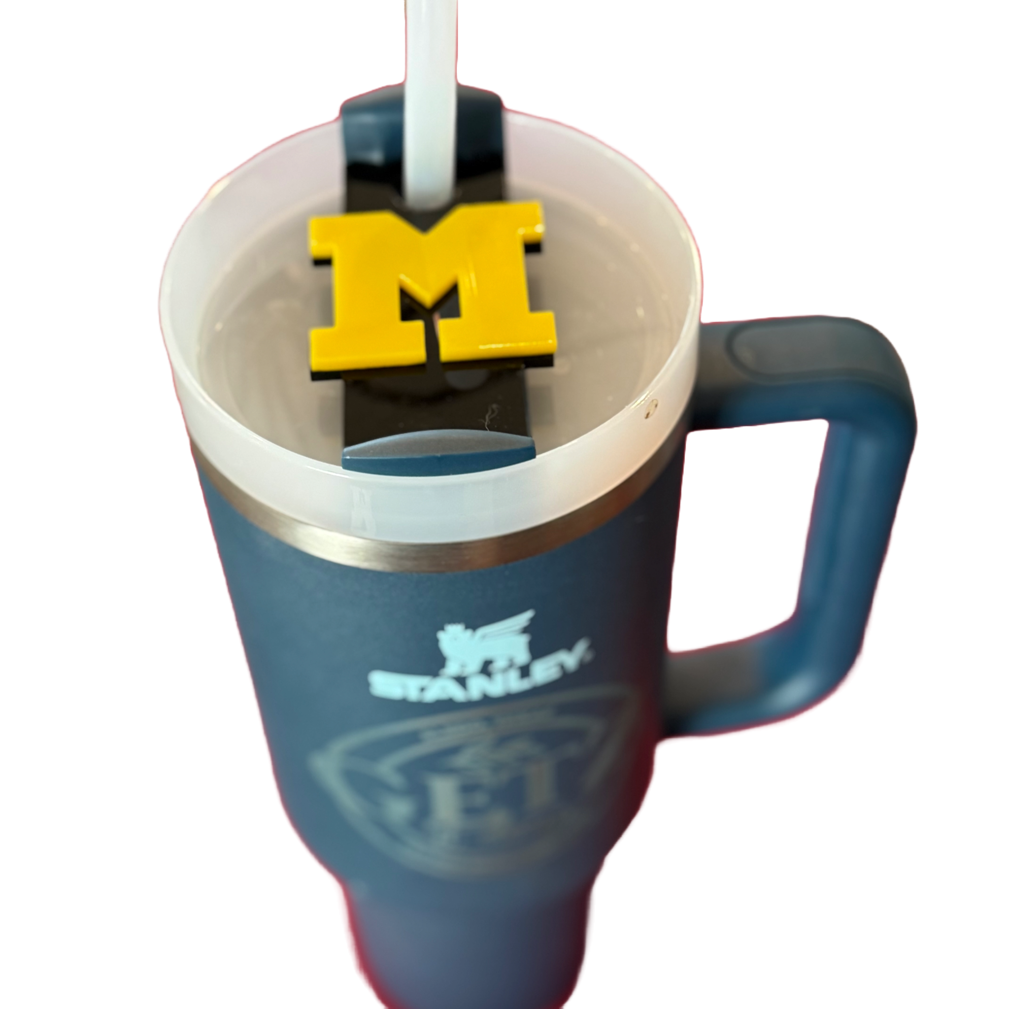 A blue Stanley travel mug with a handle, featuring the embossed brand logo. Inside, a straw leads through a yellow cube-shaped object with a prominent letter M on top, floating on the beverage. The background is white and uncluttered.