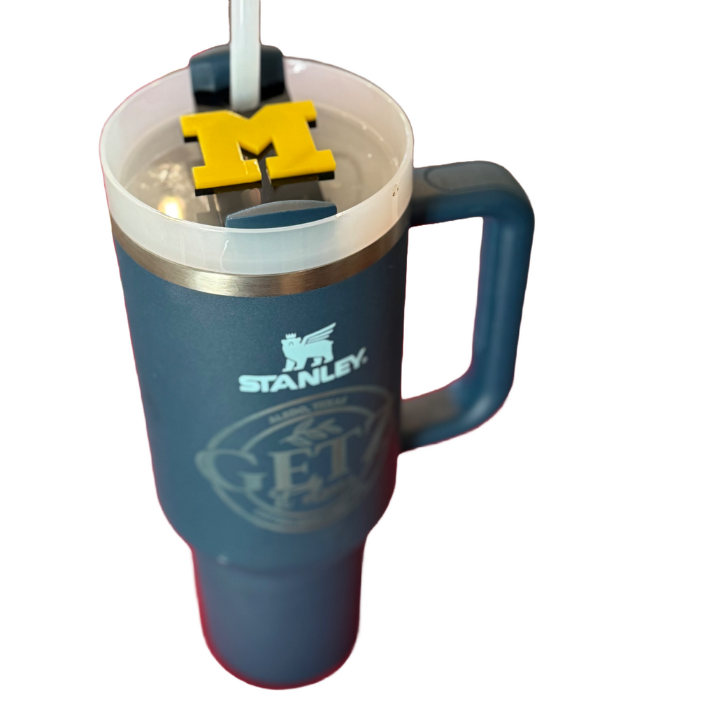 A large blue Stanley mug with a handle and a clear lid featuring a yellow M logo decoration. The mug has a distinct logo on its side. The straw is white, extending through the lid. The background is plain white, highlighting the mug.