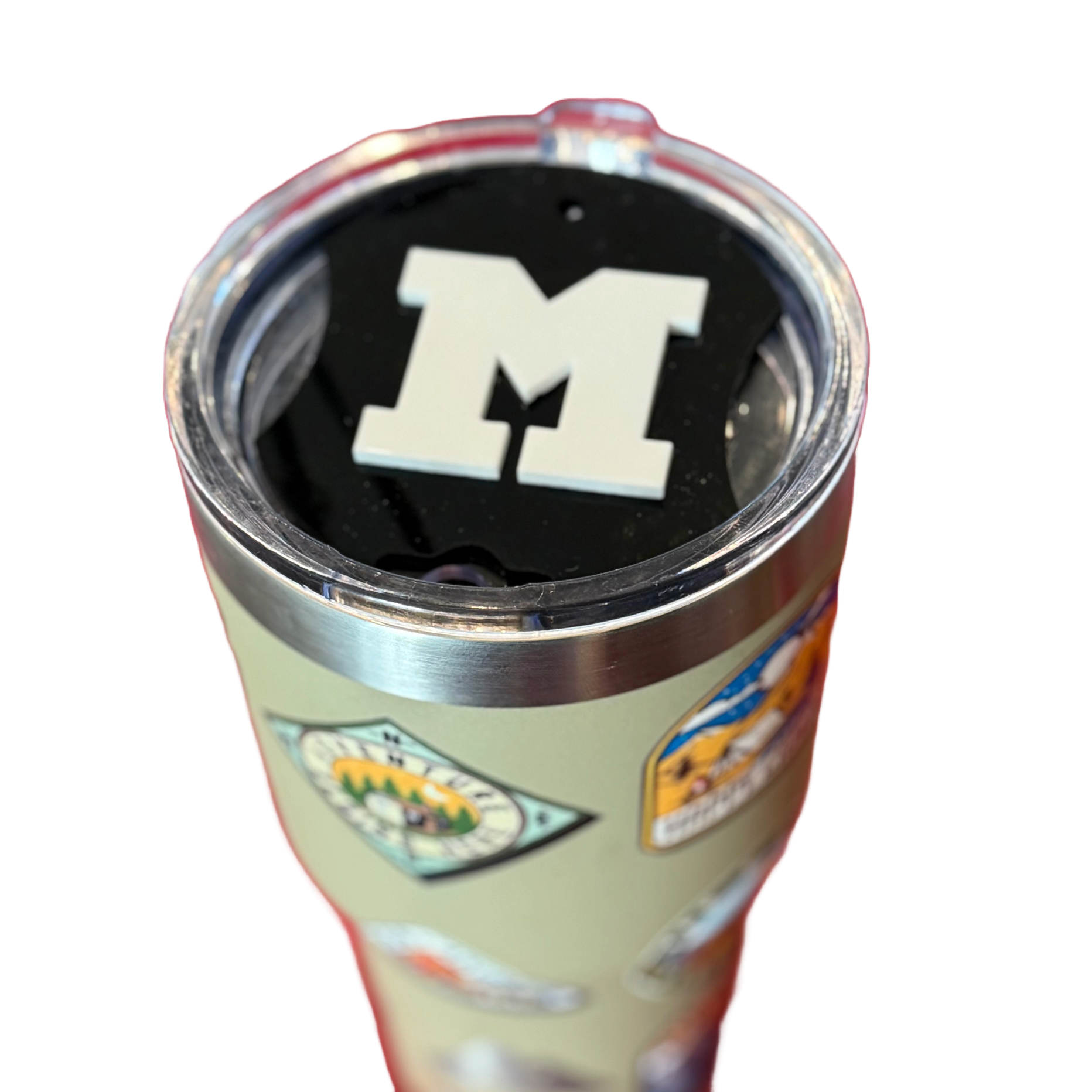 A travel tumbler with a clear lid featuring a bold, white letter M on top. The tumbler is covered with various colorful stickers depicting landscapes and logos, suggesting travel or outdoor themes. The background is white, highlighting the tumblers design.