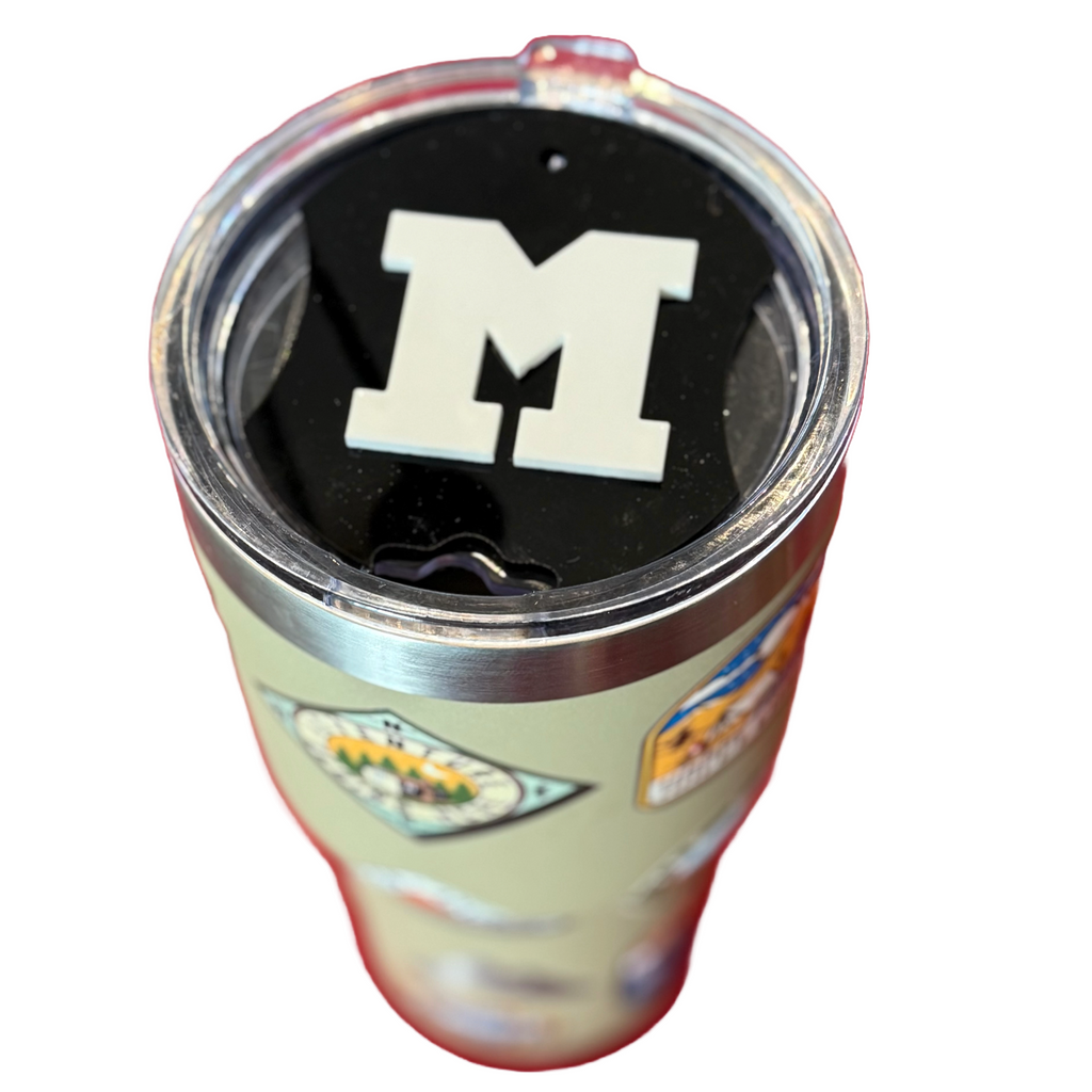 A stainless steel tumbler with various colorful stickers sits on a surface. The lid features a large, bold white letter M on a black background. The stickers depict different outdoor-themed designs, including mountains and badges. The tumbler is isolated on a white background.