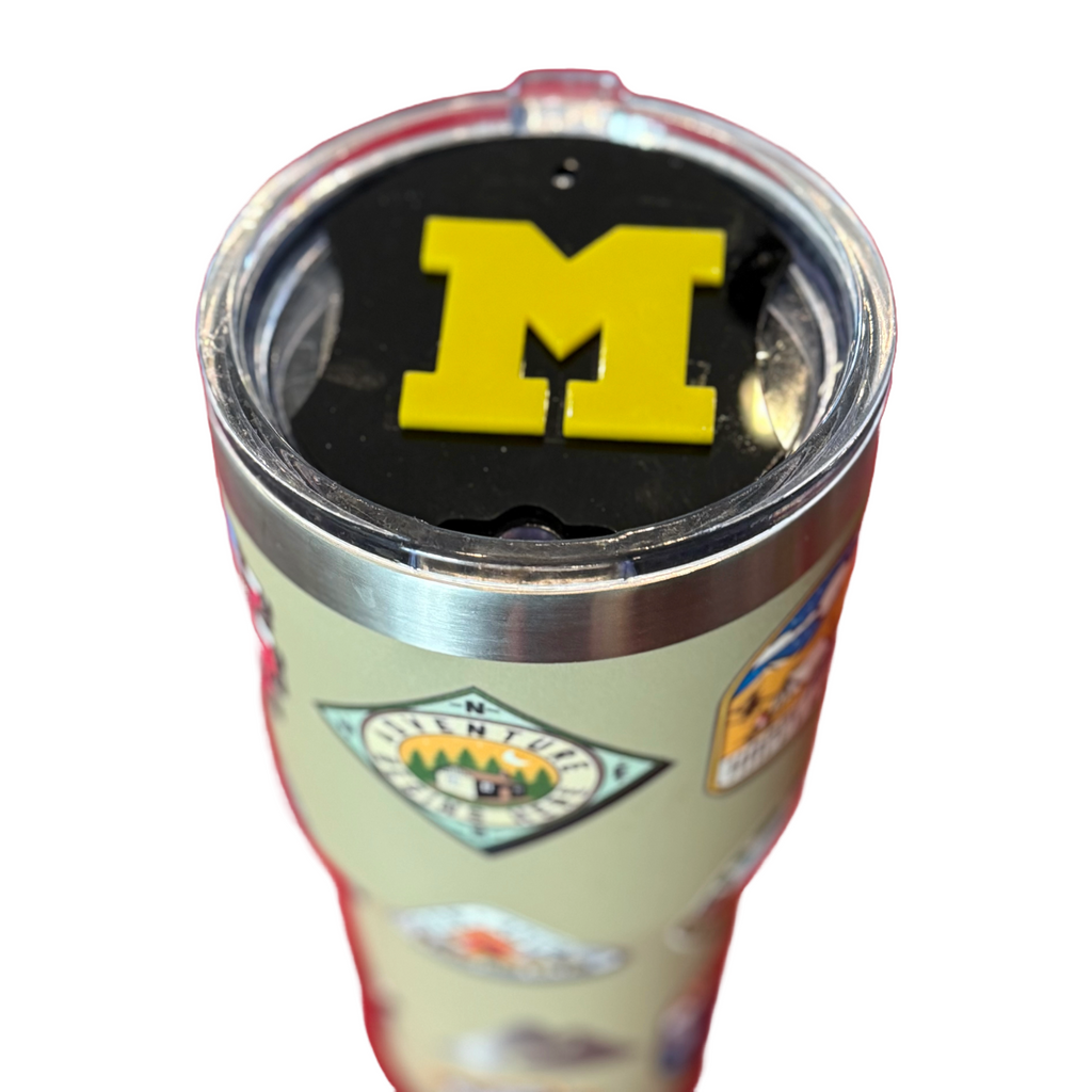 A silver tumbler with logos and stickers is topped with a black lid featuring a bold, yellow M emblem. The background is white, emphasizing the tumblers design and vibrant colors.