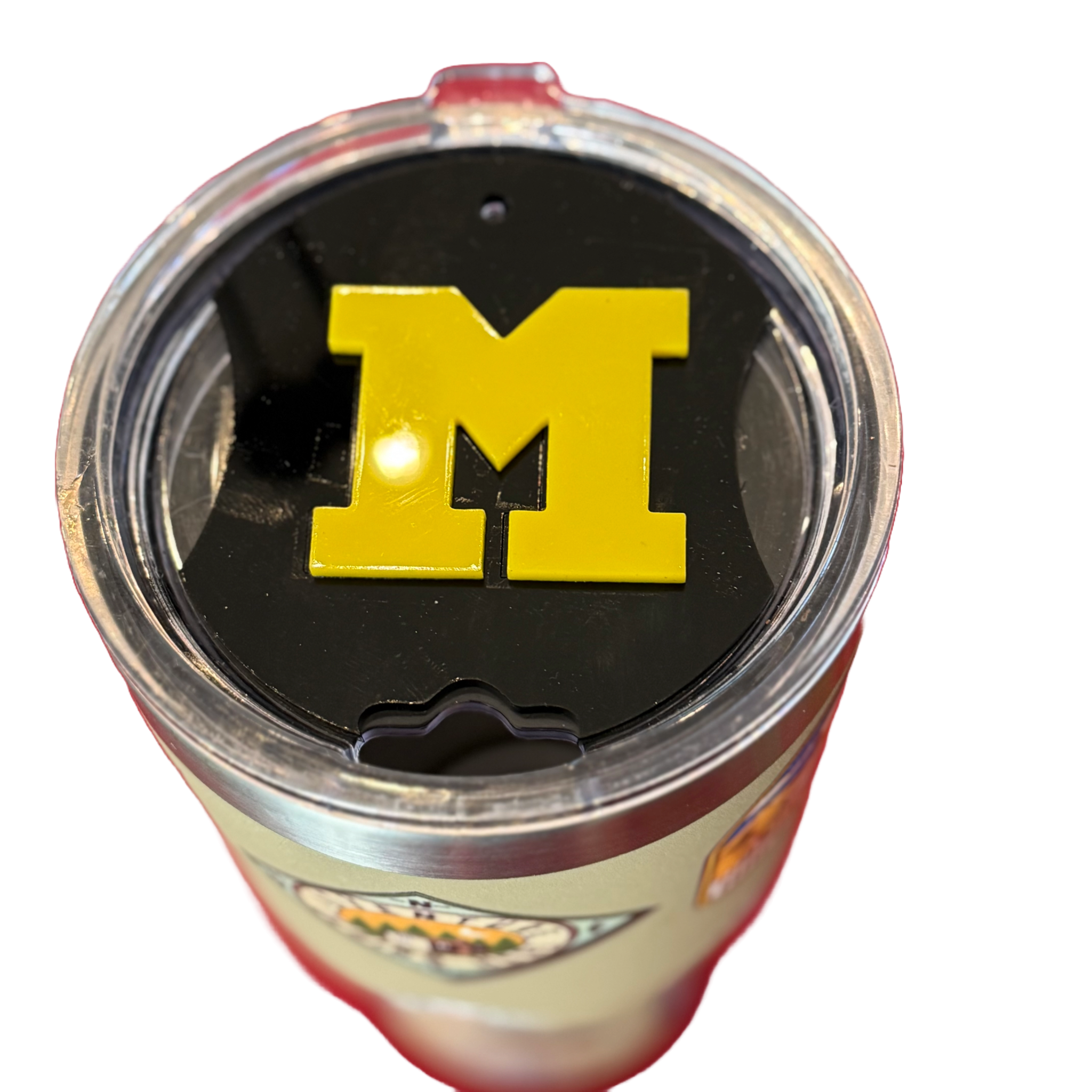 A close-up of a tumbler with a transparent lid featuring a prominent yellow M logo on a black background. The tumbler has a silver base and includes colorful logo designs on its sides. The image is taken from above on a white background.