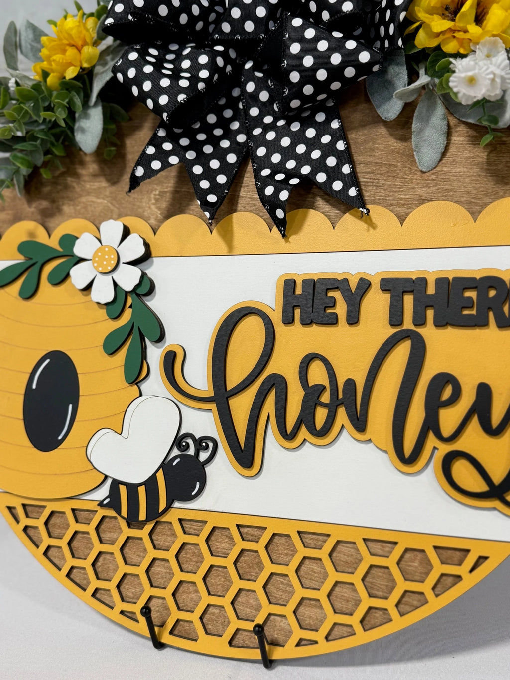 A close-up of a decorative sign featuring a bee theme. It has a yellow and black cartoon bee, honeycomb pattern, and the text Hey there honey in bold, playful font. The sign is adorned with a polka dot bow, white flowers, and green leaves over a wooden background.