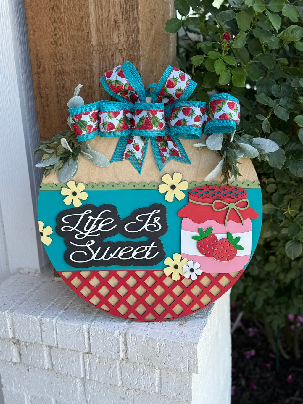 A round decorative sign with a bow, flowers, and jars of jam features the phrase Life Is Sweet in white script. The design includes strawberries and lattice patterns, and is displayed outdoors near a plant.