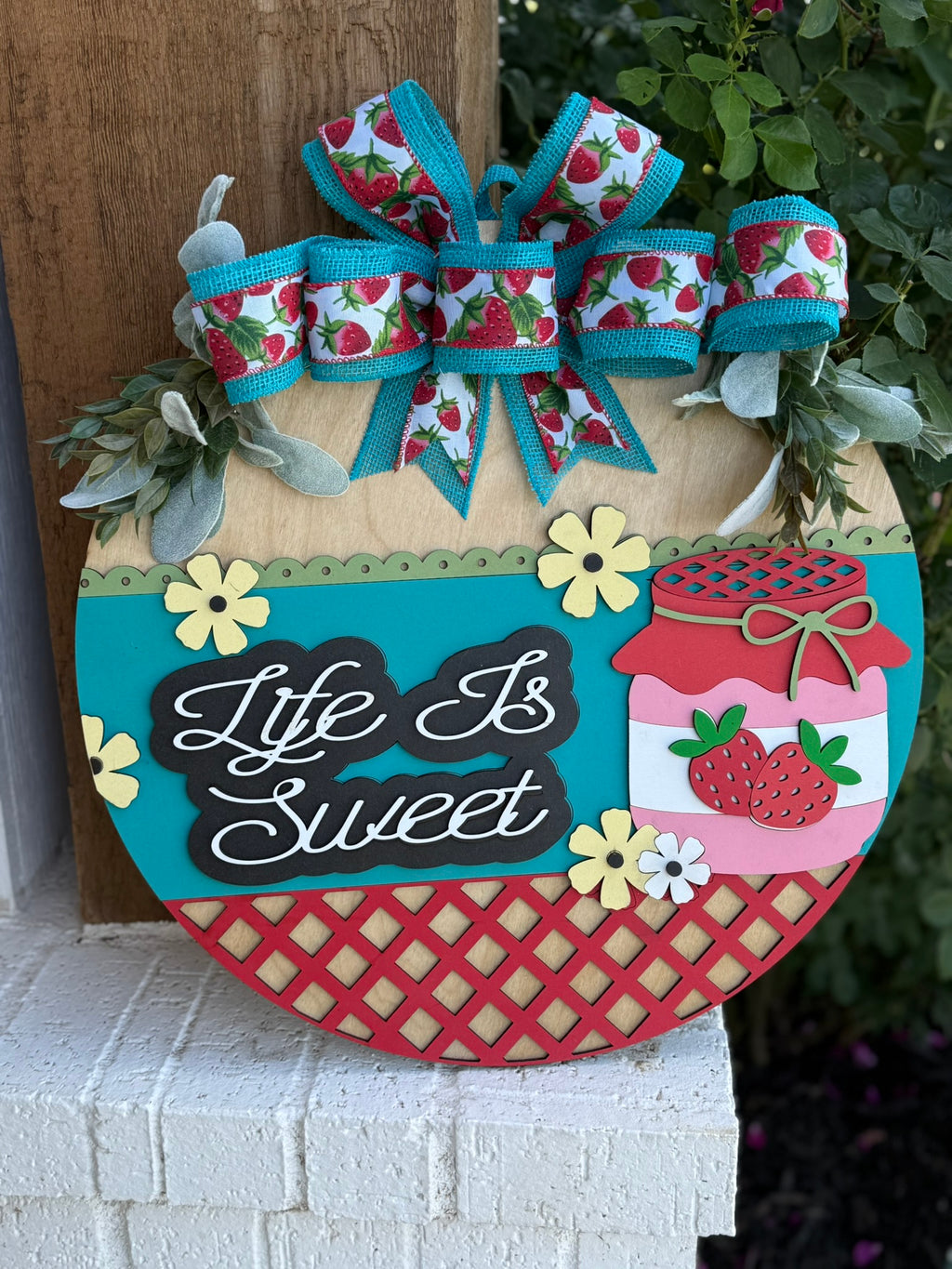 A colorful wooden sign decorated with bows and strawberry-themed ribbons, flowers, and a jar of strawberry jam reads Life Is Sweet in white script. The sign rests against a post with green leaves above it.