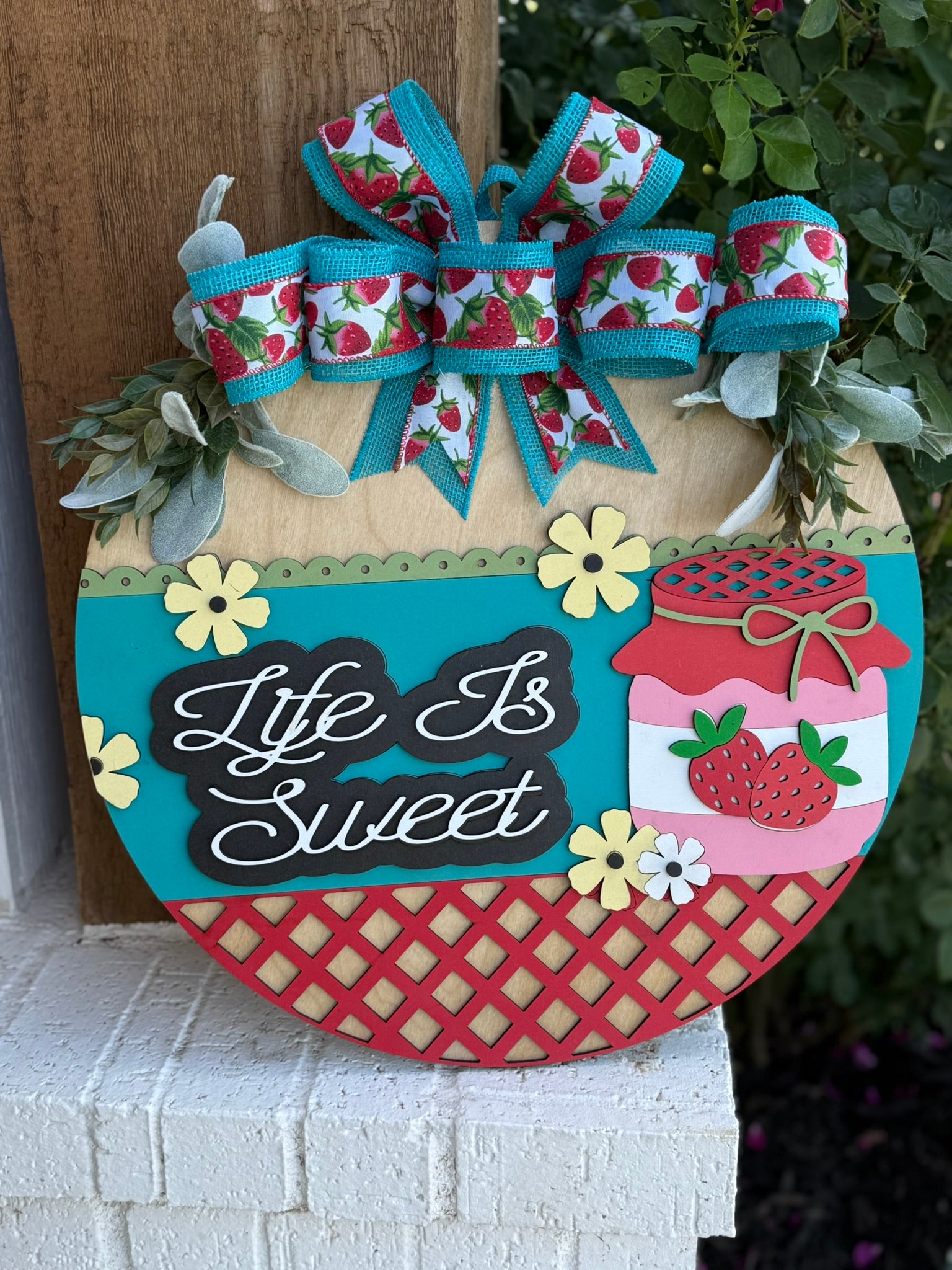 A colorful wooden sign decorated with bows and strawberry-themed ribbons, flowers, and a jar of strawberry jam reads Life Is Sweet in white script. The sign rests against a post with green leaves above it.