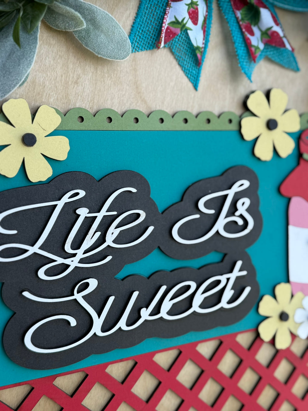 A colorful decorative sign with the words Life Is Sweet in white script on a black background, surrounded by paper flowers and a red lattice design, with teal, yellow, and floral accents.