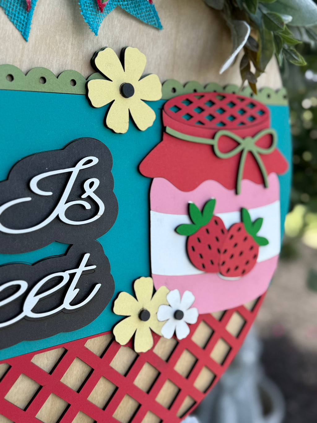 A close-up of a colorful decorative sign featuring paper cutouts of flowers, strawberries, and a jar of jam on a teal and red lattice background. The partial words Is sweet are also visible.