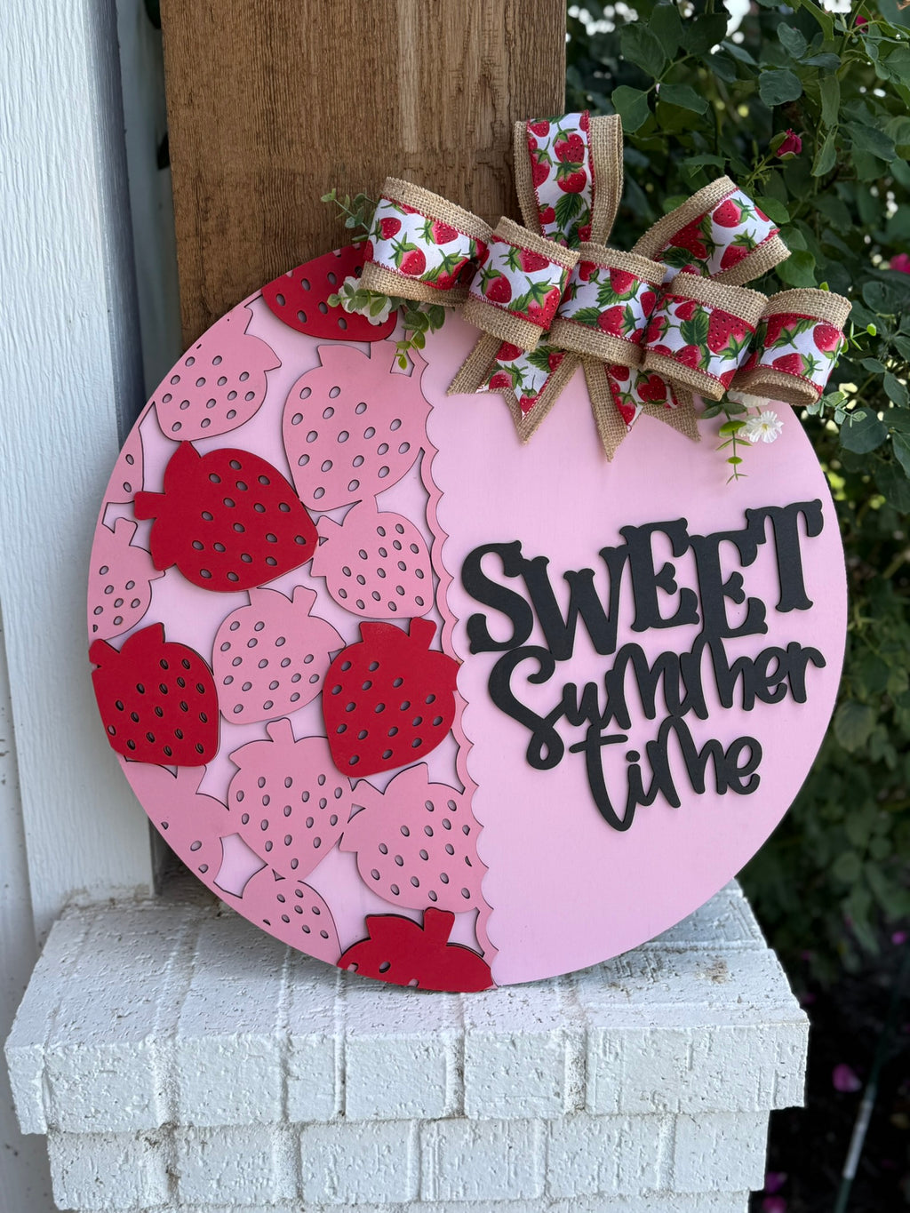 A round pink wooden sign decorated with red and pink strawberries, a large ribbon with strawberry designs, and the words “SWEET Summer time” in black letters, leaning against a porch post next to green leaves.