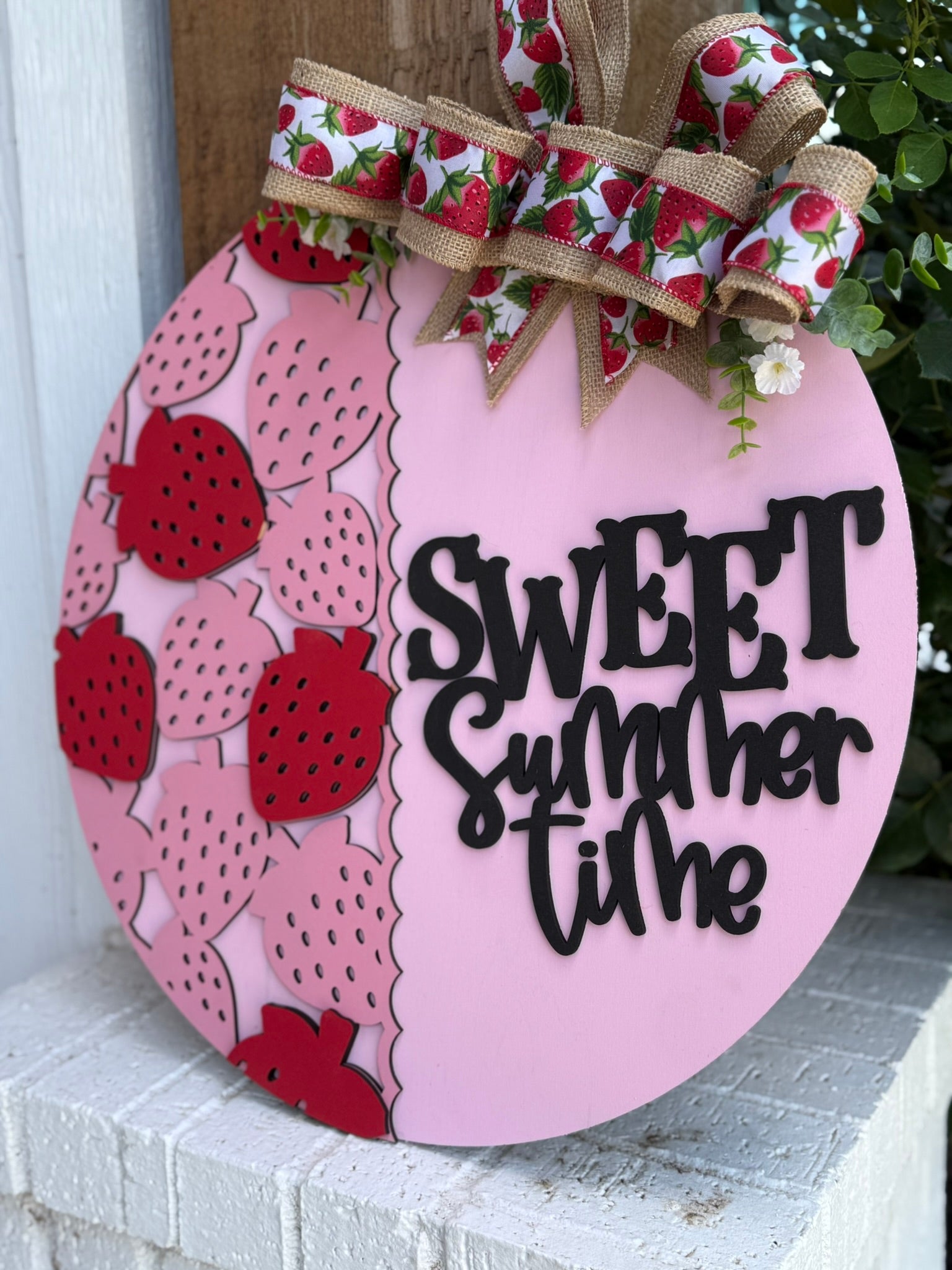 A round pink sign decorated with red strawberries, a scalloped edge, a large bow with strawberry-patterned ribbon, and the words Sweet Summer Time in bold black letters. The sign is displayed on a white brick surface.