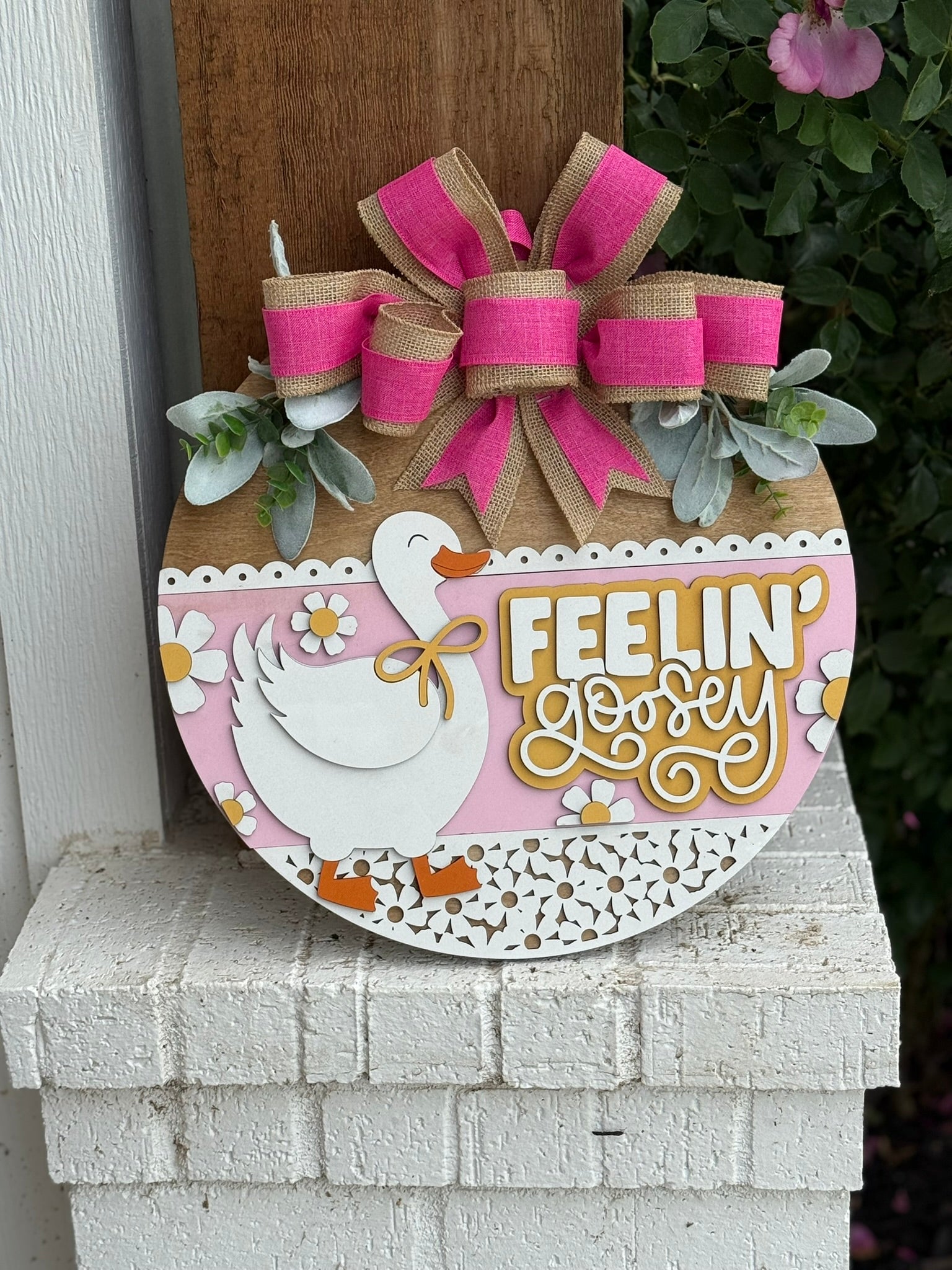 A round decorative sign with a white goose, daisies, and the text FEELIN GOOSEY on a pink background, topped with a large pink and burlap bow and greenery, hanging on a white brick wall.