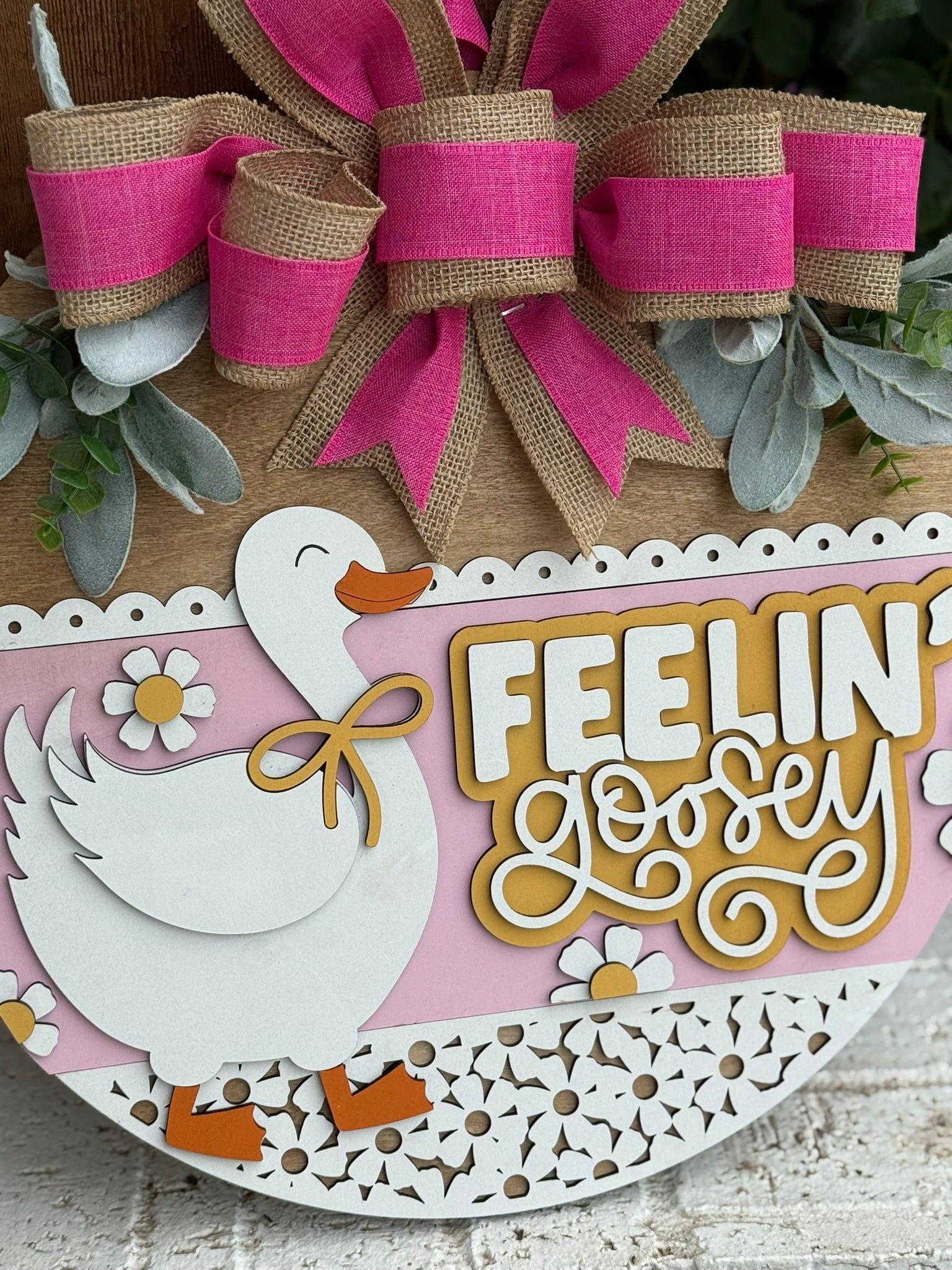 A decorative sign features a smiling white goose with yellow feet, surrounded by flowers and greenery. Above the goose, a pink and burlap bow is tied. The sign reads, FEELIN’ goosey in yellow and white letters.