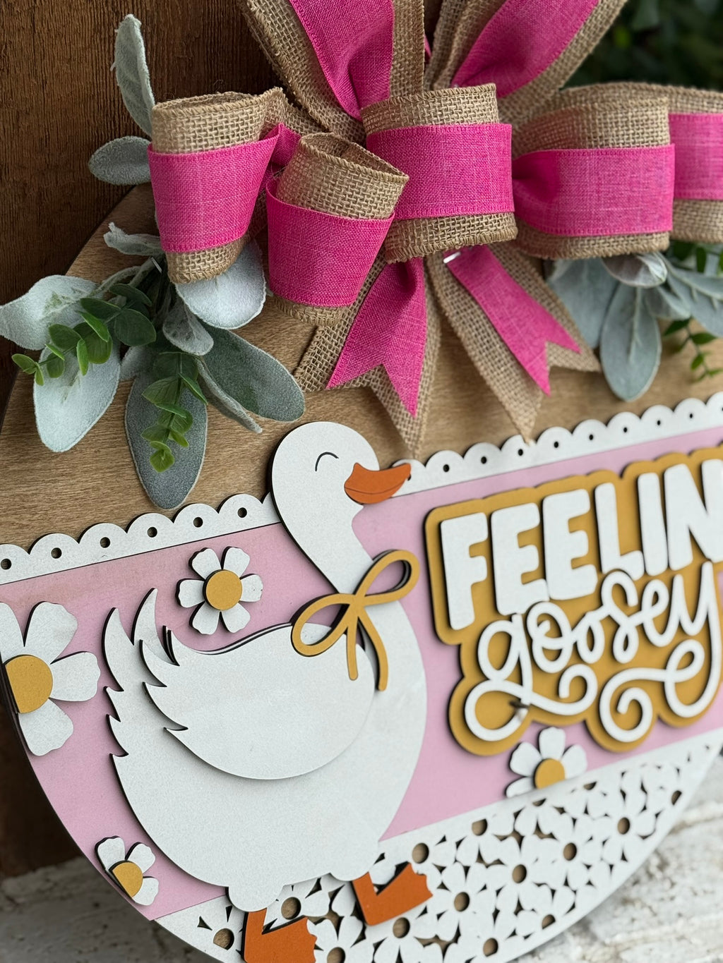 A decorative sign with a white duck, yellow beak, and bow tie, surrounded by flowers and greenery, featuring a large pink and burlap bow and the phrase Feelin Goosey in bold yellow and white letters.