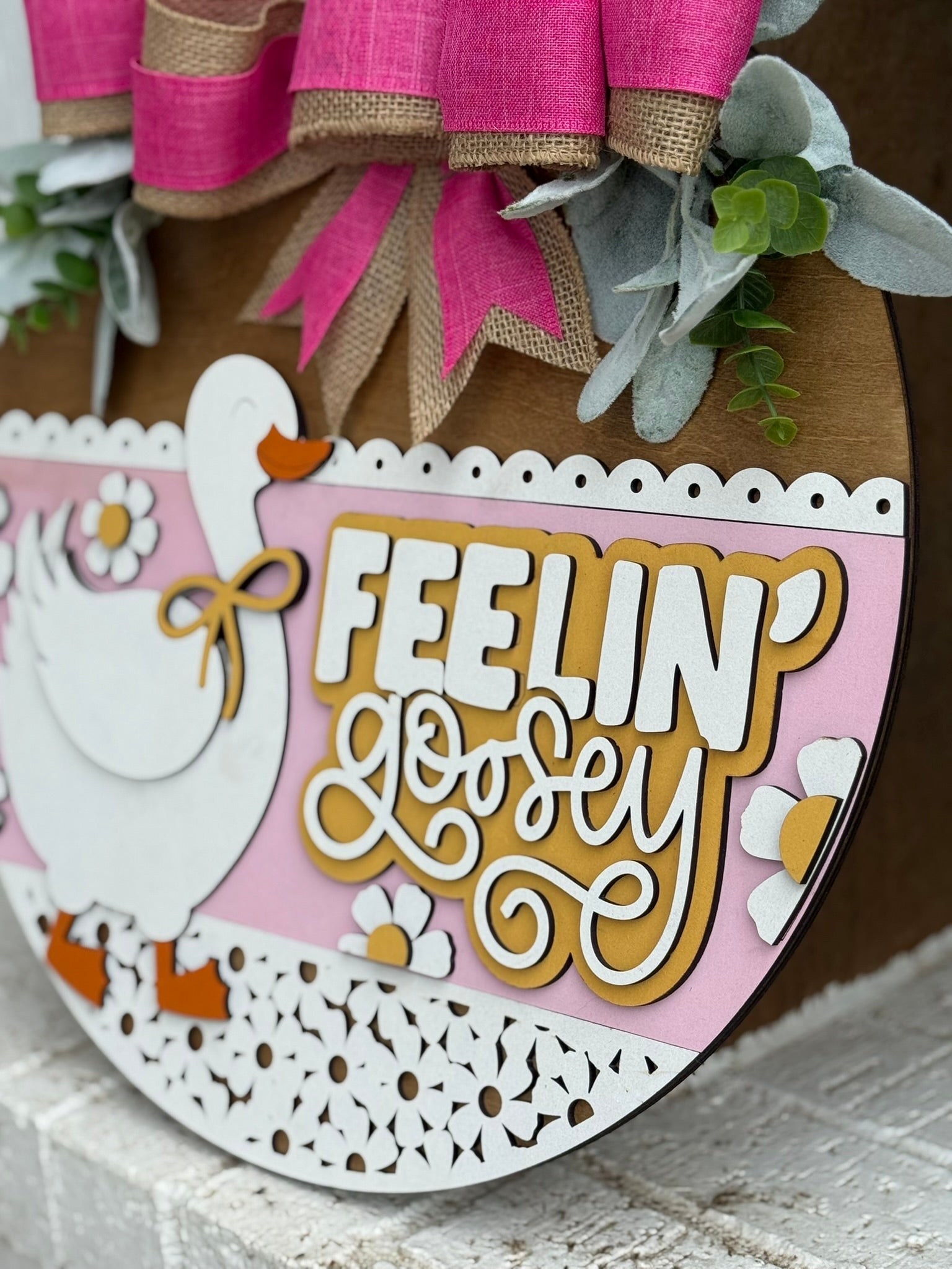 A round decorative sign features a white goose, daisies, and the phrase Feelin Goosey in layered lettering on a pink background, topped with pink and burlap ribbons and green leaves.