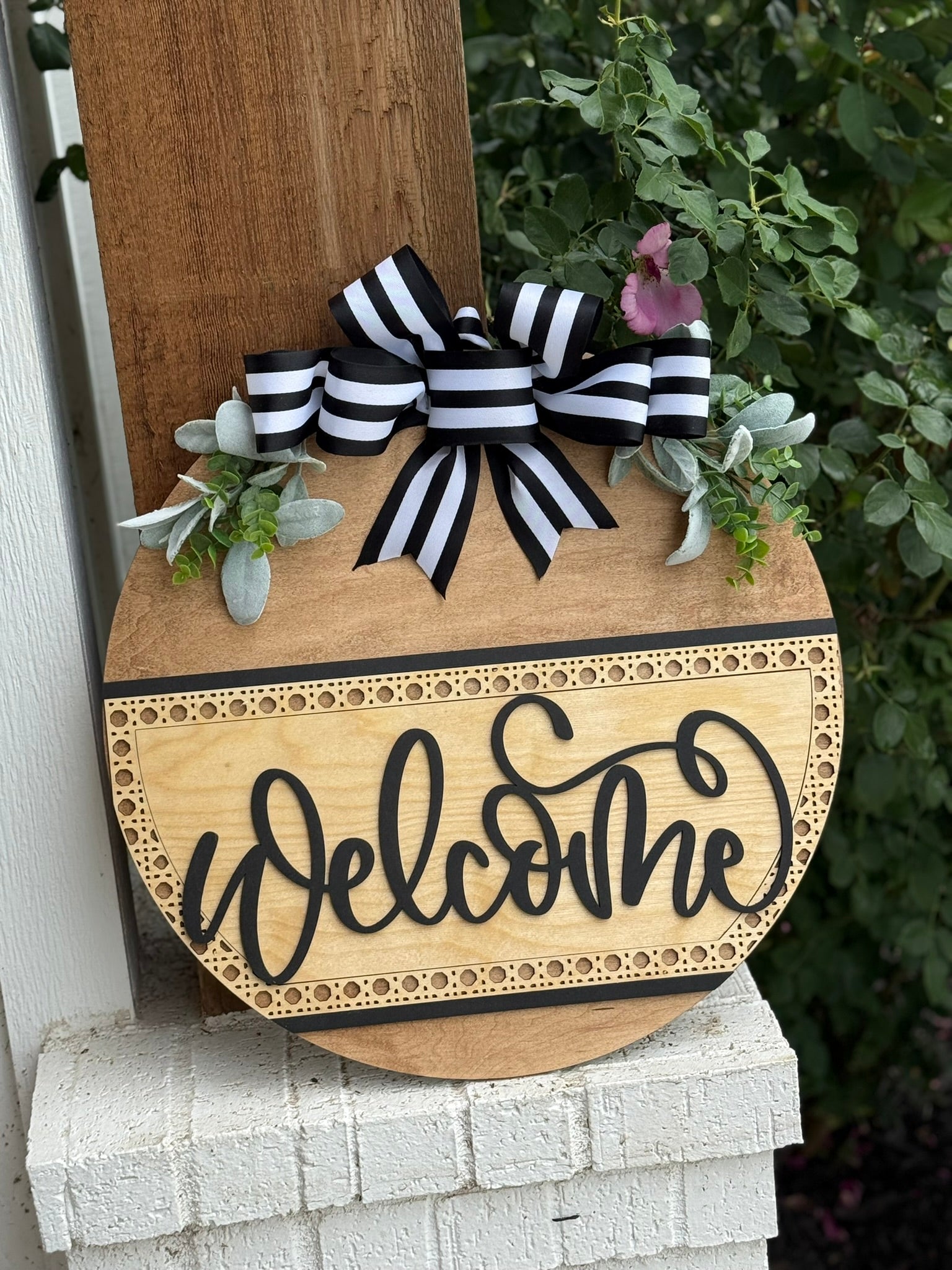 A round wooden Welcome sign hangs on a porch post, decorated with a black-and-white striped bow and green foliage, with some pink flowers in the background.
