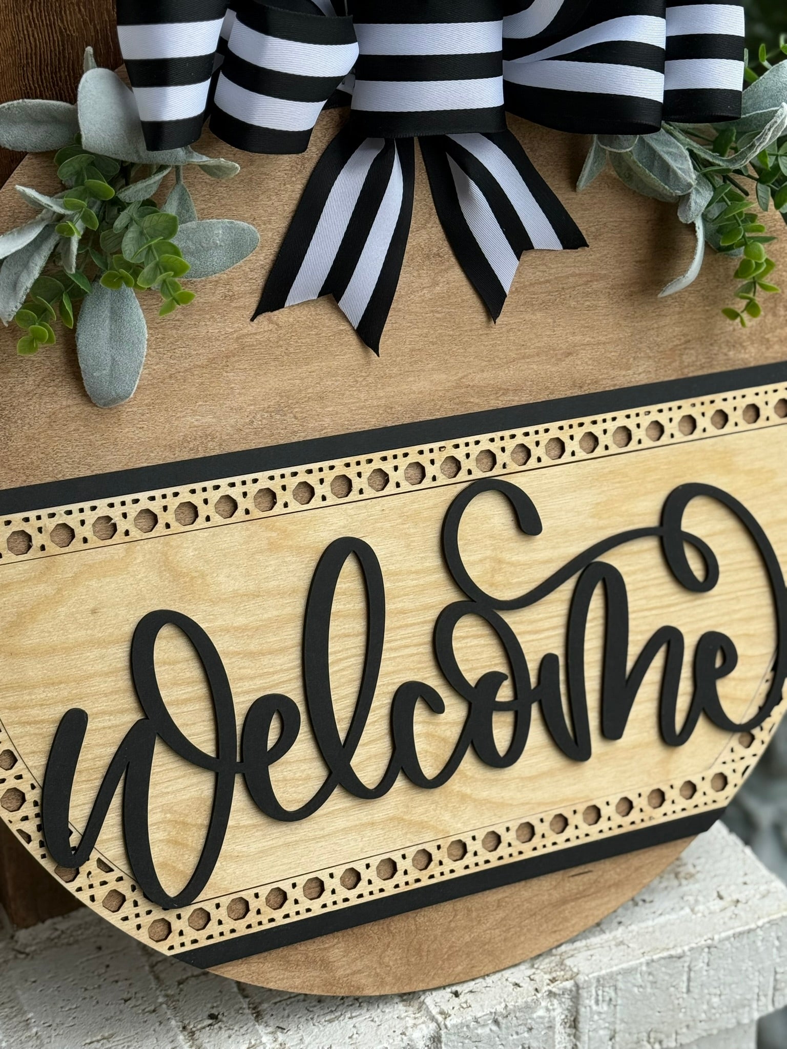 A wooden sign decorated with greenery and a black-and-white striped bow displays the word Welcome in large, cursive black letters. The sign has a decorative border and rests on a white brick surface.