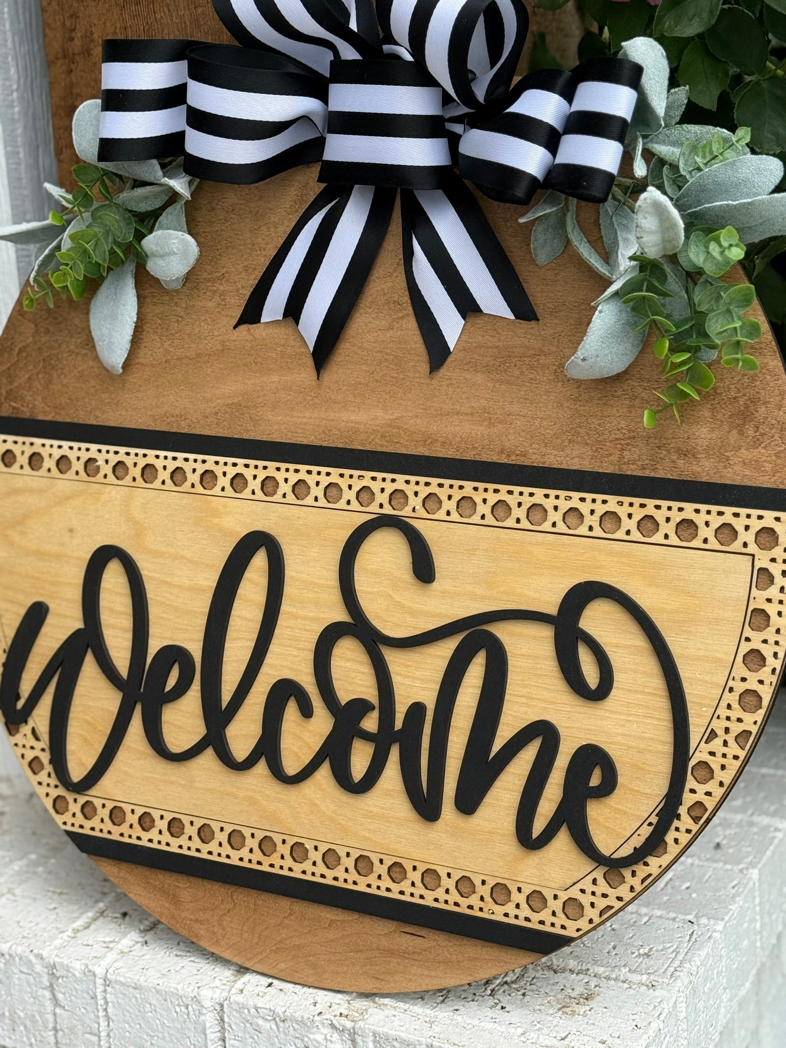 A round wooden sign with the word welcome in black script, decorated with green leaves and a black-and-white striped bow at the top, is displayed against a white brick background.
