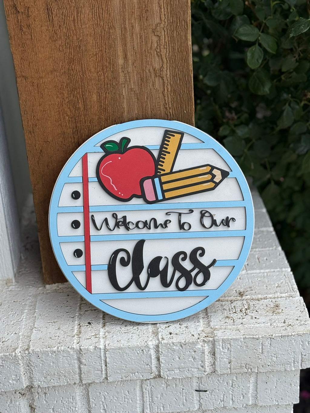 A round sign with a red apple, two pencils, and lined paper design reads “Welcome to Our Class.” It is propped against a wooden post on a white brick surface, with green foliage in the background.