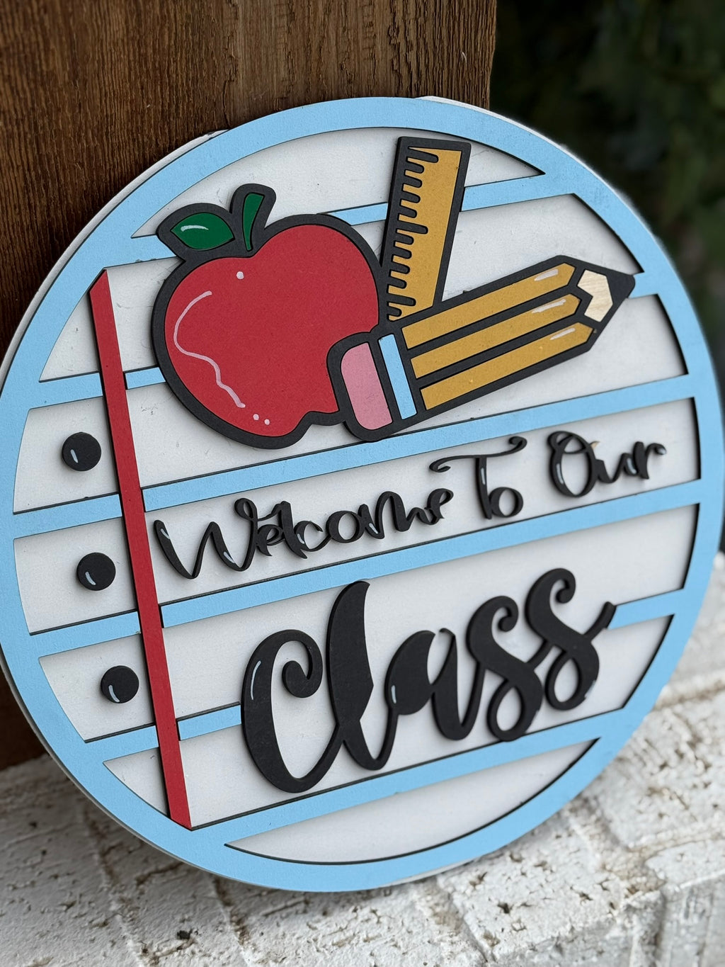 A circular sign with a red apple, yellow pencil, and ruler, styled like lined notebook paper, reads Welcome To Our Class in decorative black lettering. The sign is leaning against a wooden surface.