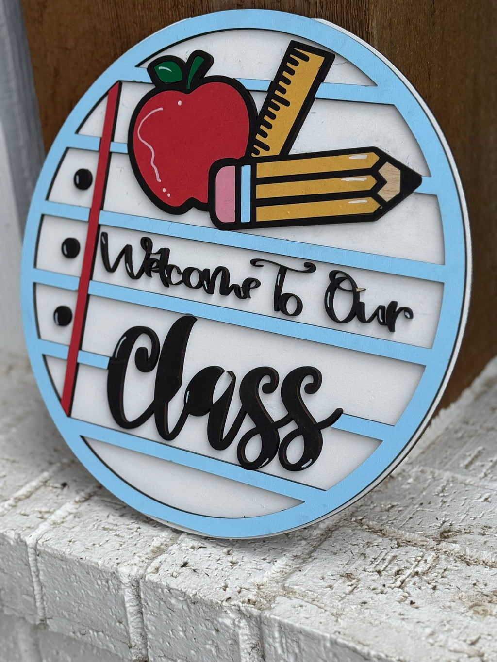 Round sign with light blue trim, a red apple, yellow pencil, and notebook paper lines. The text reads Welcome To Our Class in black script and bold letters. The sign is propped against a wooden surface.