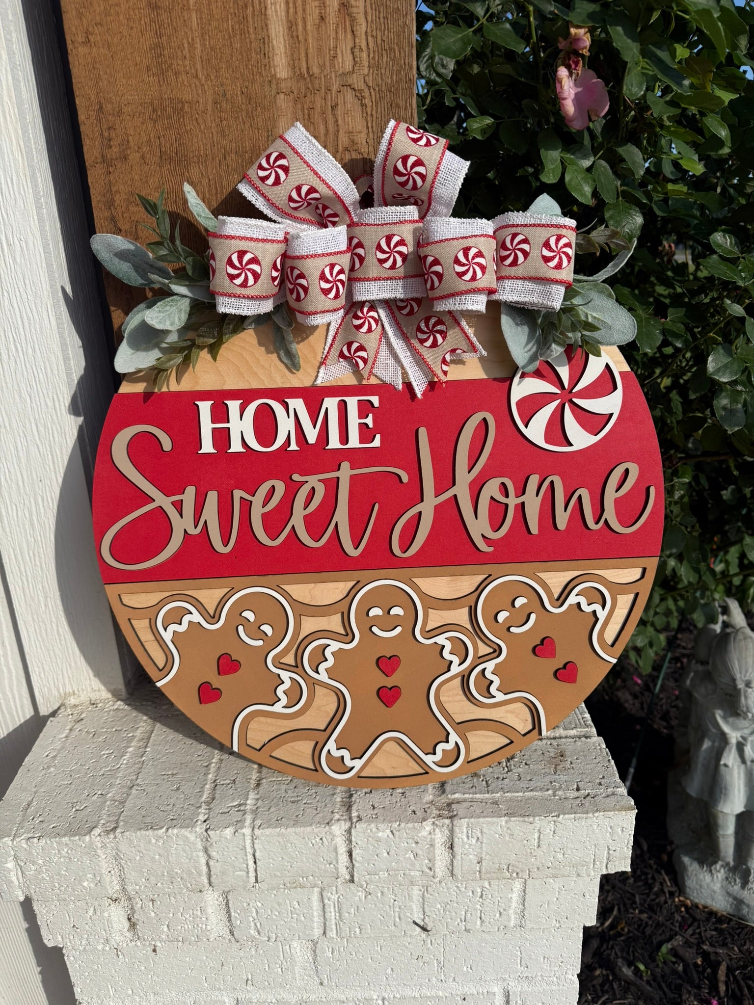 Round holiday sign with Home Sweet Home text, decorated with ribbon, faux greenery, peppermint candy designs, and three smiling gingerbread figures with red hearts, displayed outdoors against a brick ledge and foliage.