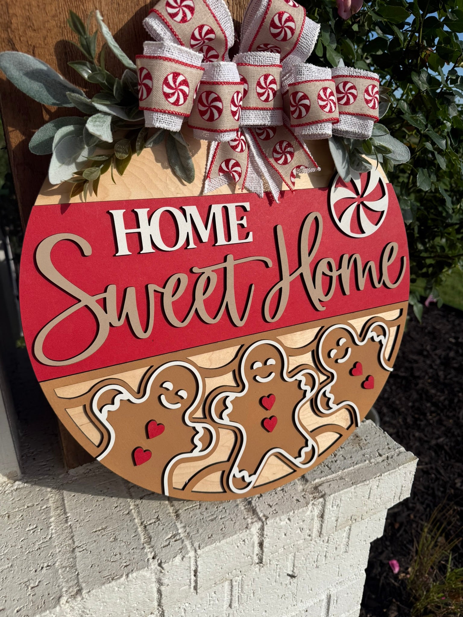 A round wooden sign decorated with ribbon and faux greenery, featuring the words Home Sweet Home with gingerbread figures and peppermint designs. The sign sits on a white brick ledge near a garden.