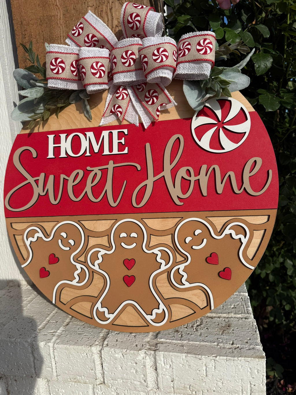 A round wooden sign decorated with gingerbread people, red hearts, and a red-and-white peppermint design. It reads Home Sweet Home and is topped with a bow featuring peppermint candy patterns.
