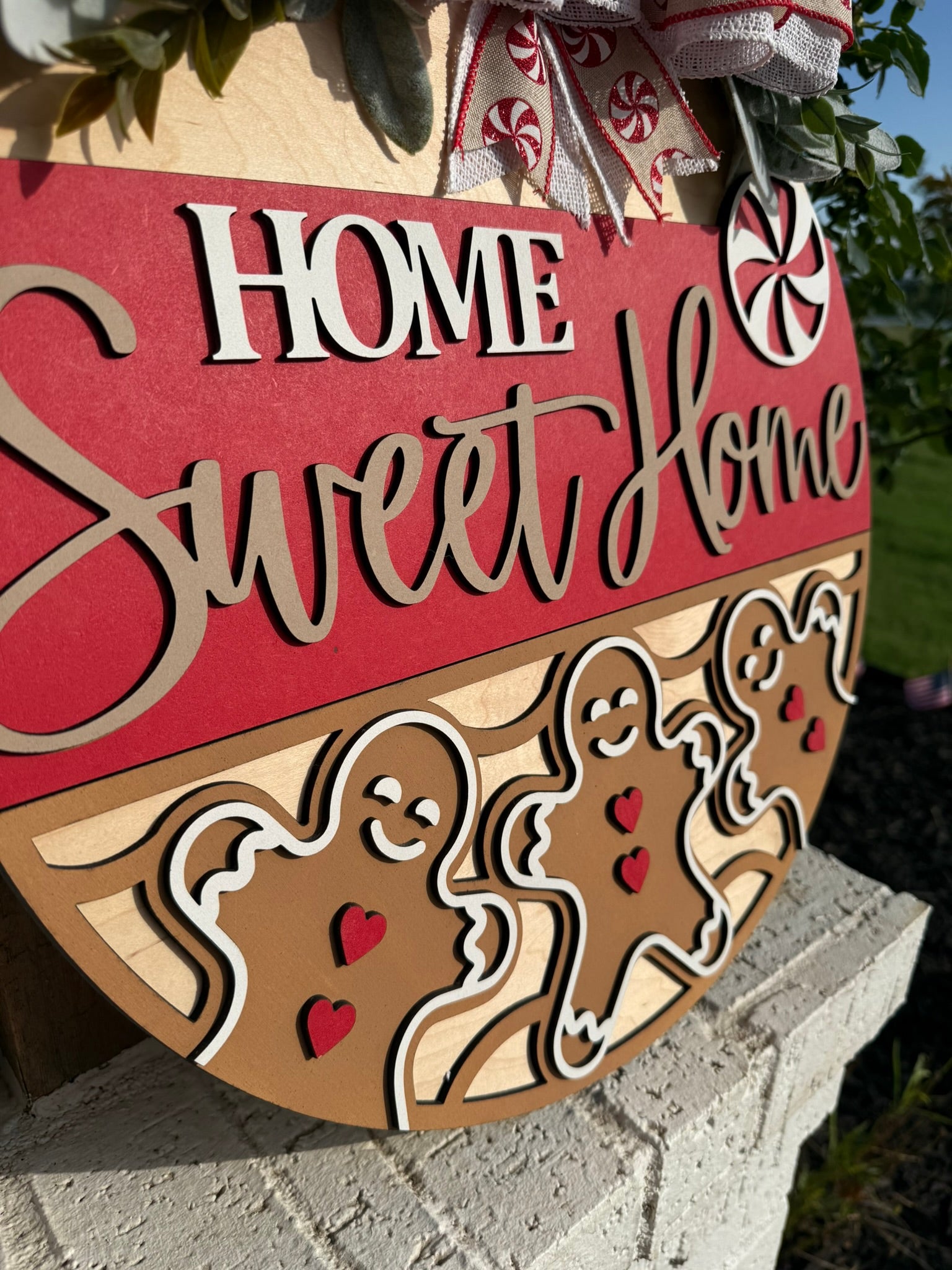 A wooden sign reads Home Sweet Home with gingerbread figures and hearts, decorated with red and white accents and peppermint candies, sitting on a white brick surface outside.