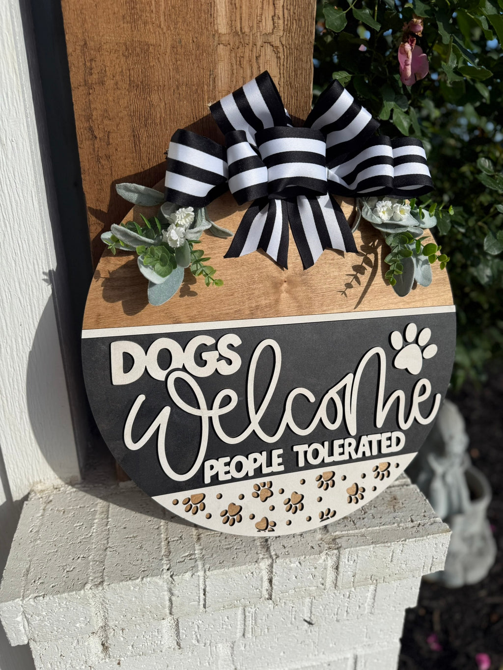 A round wooden sign with a black and white striped bow, artificial greenery, and text that reads: Dogs Welcome, People Tolerated, with paw print decorations at the bottom, hanging outside on a brick shelf.