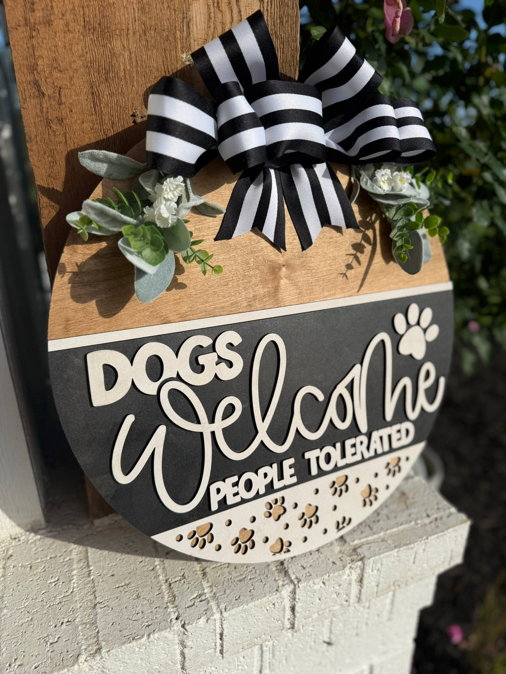 A round wooden sign with a black and white striped bow, greenery, and text that reads Dogs welcome, people tolerated, with paw prints decorating the bottom.