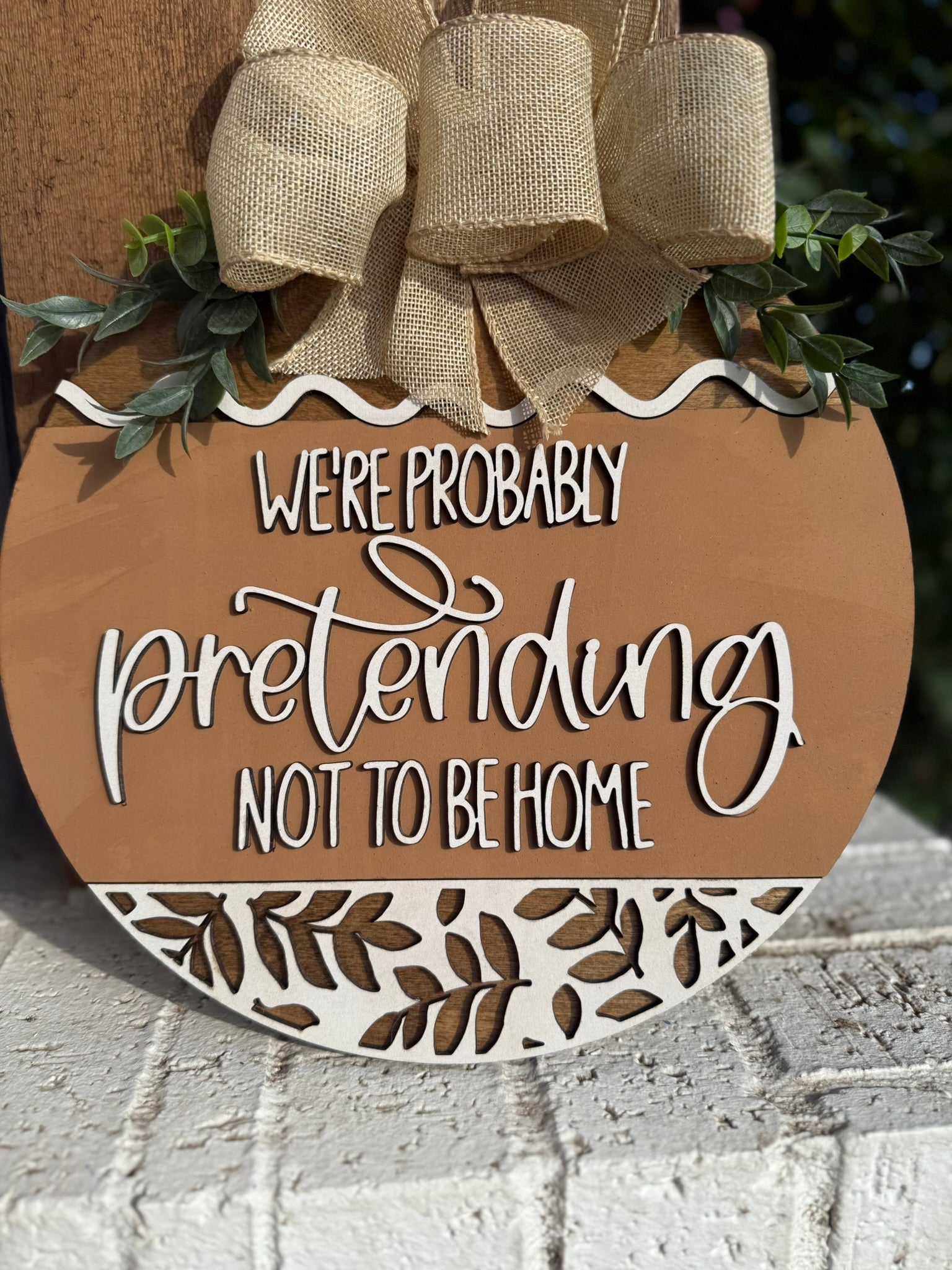 A round decorative sign with a burlap bow and greenery reads, Were probably pretending not to be home, in white script on a brown background with white leaf patterns at the bottom.