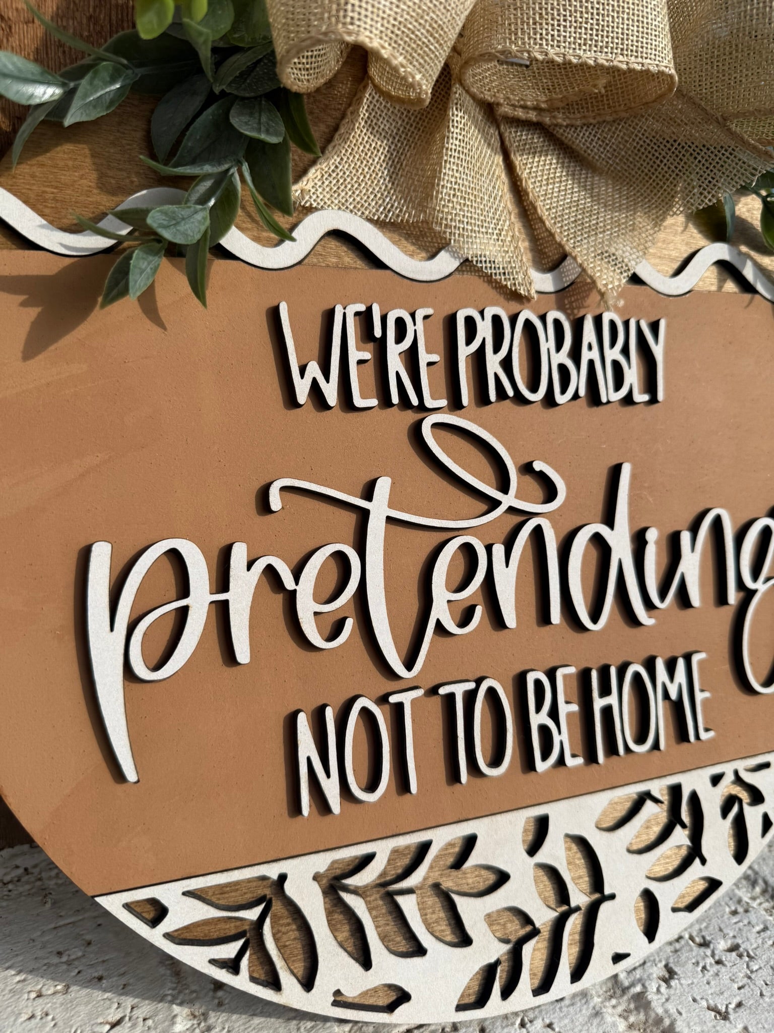 A decorative wooden sign with greenery and a burlap bow reads, Were probably pretending not to be home in white, playful script. The bottom features a carved leaf pattern.