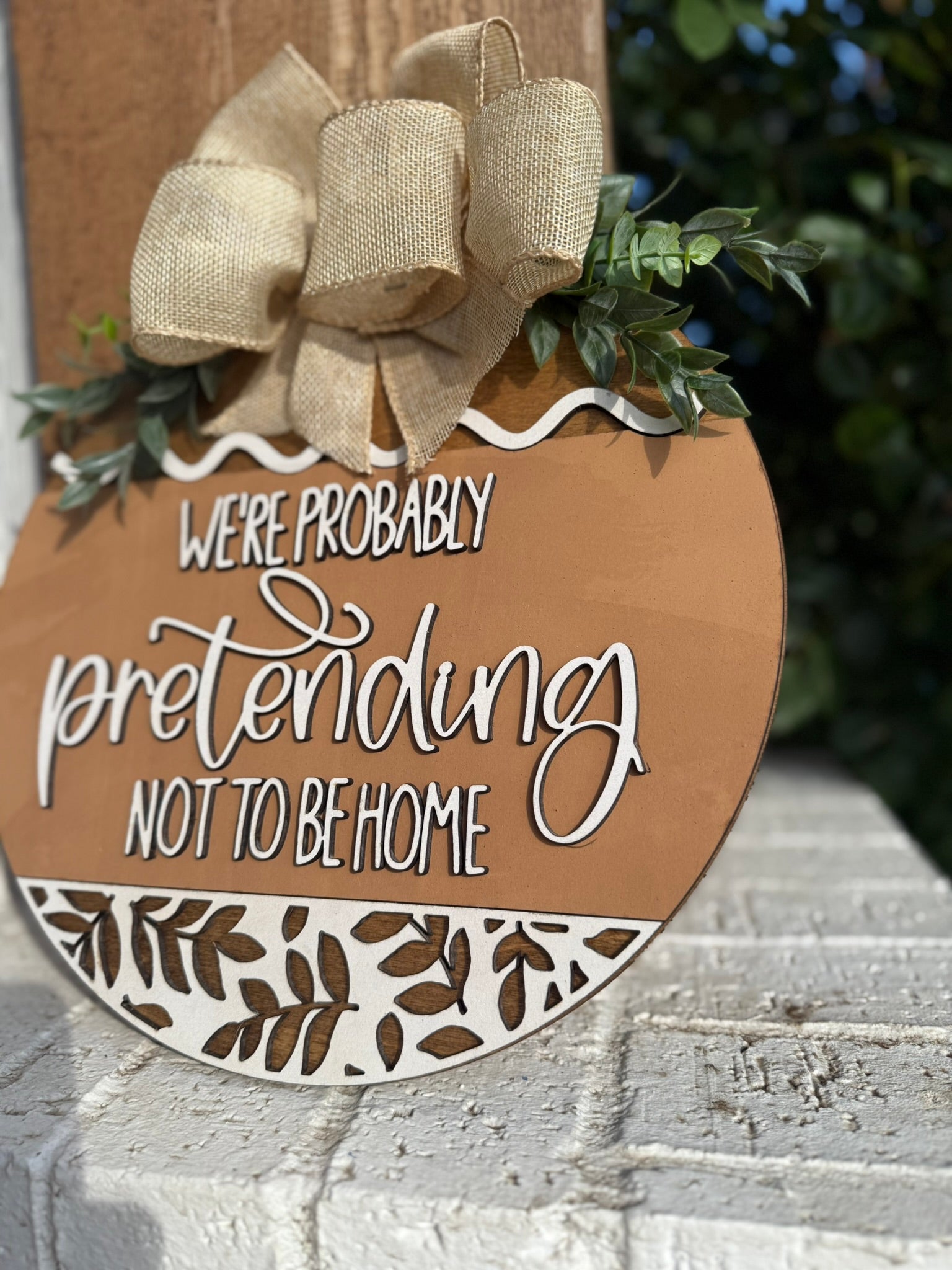 A decorative sign with a burlap bow reads, Were probably pretending not to be home, and is displayed outdoors against a white brick surface with greenery in the background.