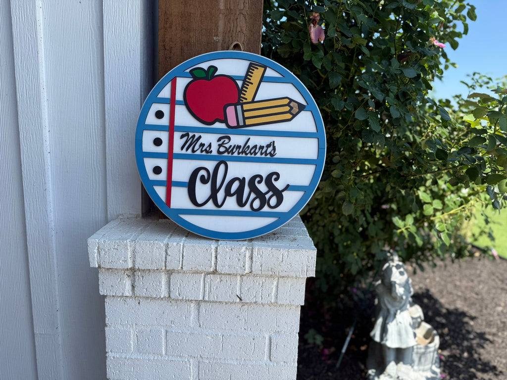 A round sign with a red apple, two yellow pencils, and the text “Mrs. Burkarts Class” rests on a white brick column outdoors, with bushes and a small garden statue in the background.