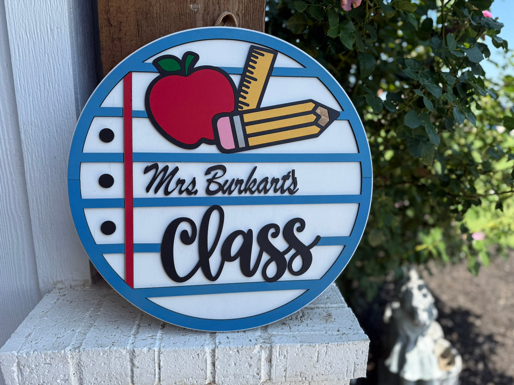 A round sign with blue and white stripes, featuring an apple, pencil, and ruler, and text that reads “Mrs. Burkart’s Class.” The sign hangs outside on a white brick wall near greenery.