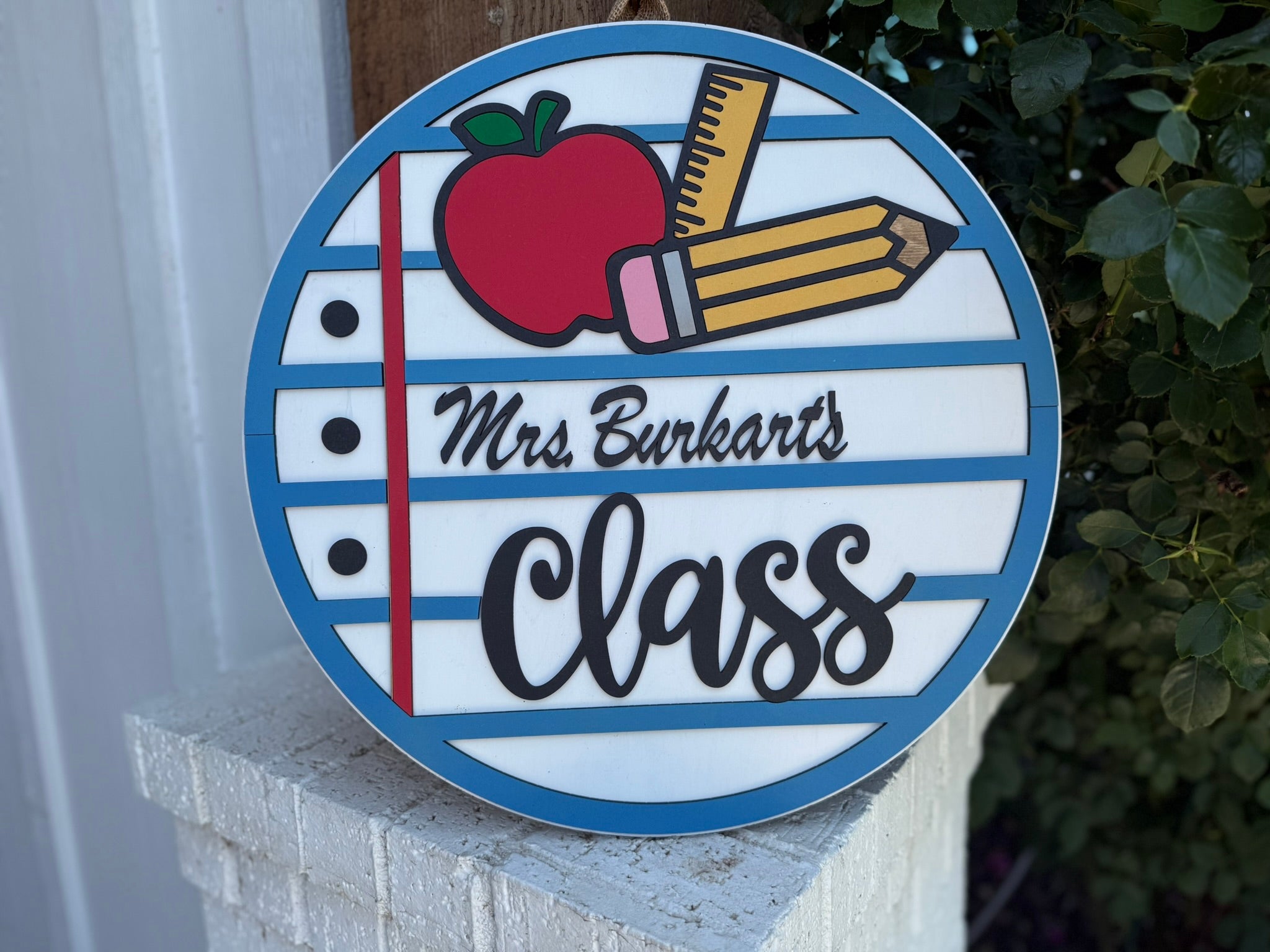 A round sign with blue and white stripes reads Mrs. Burkarts Class in script and bold letters. It features an apple, pencil, and ruler, resembling notebook paper. The sign is propped outside near green bushes.