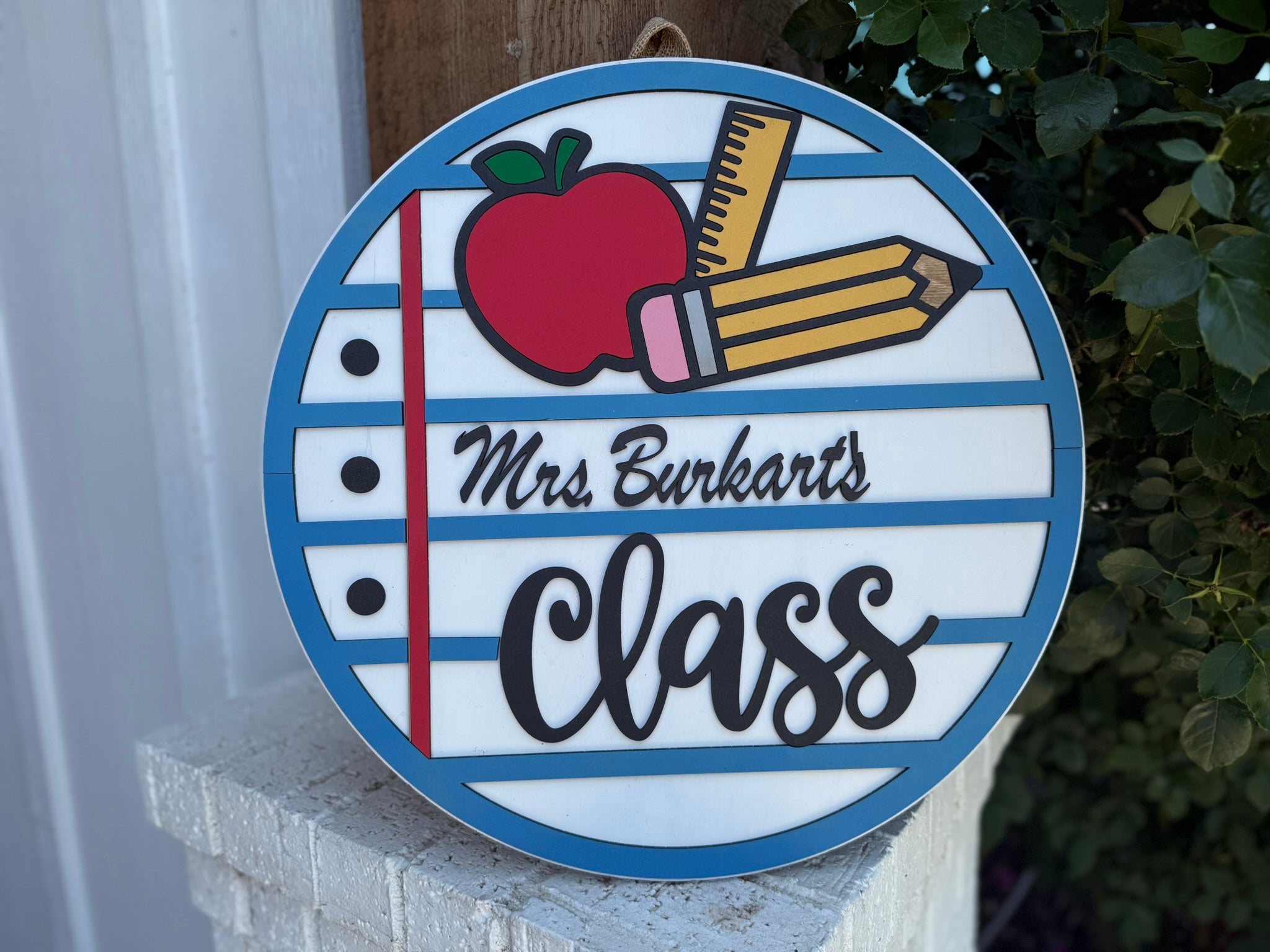 A round sign designed like notebook paper with an apple, ruler, and pencil illustration. It reads Mrs. Burkharts Class in bold and script fonts, hanging near a brick surface and leafy plant.