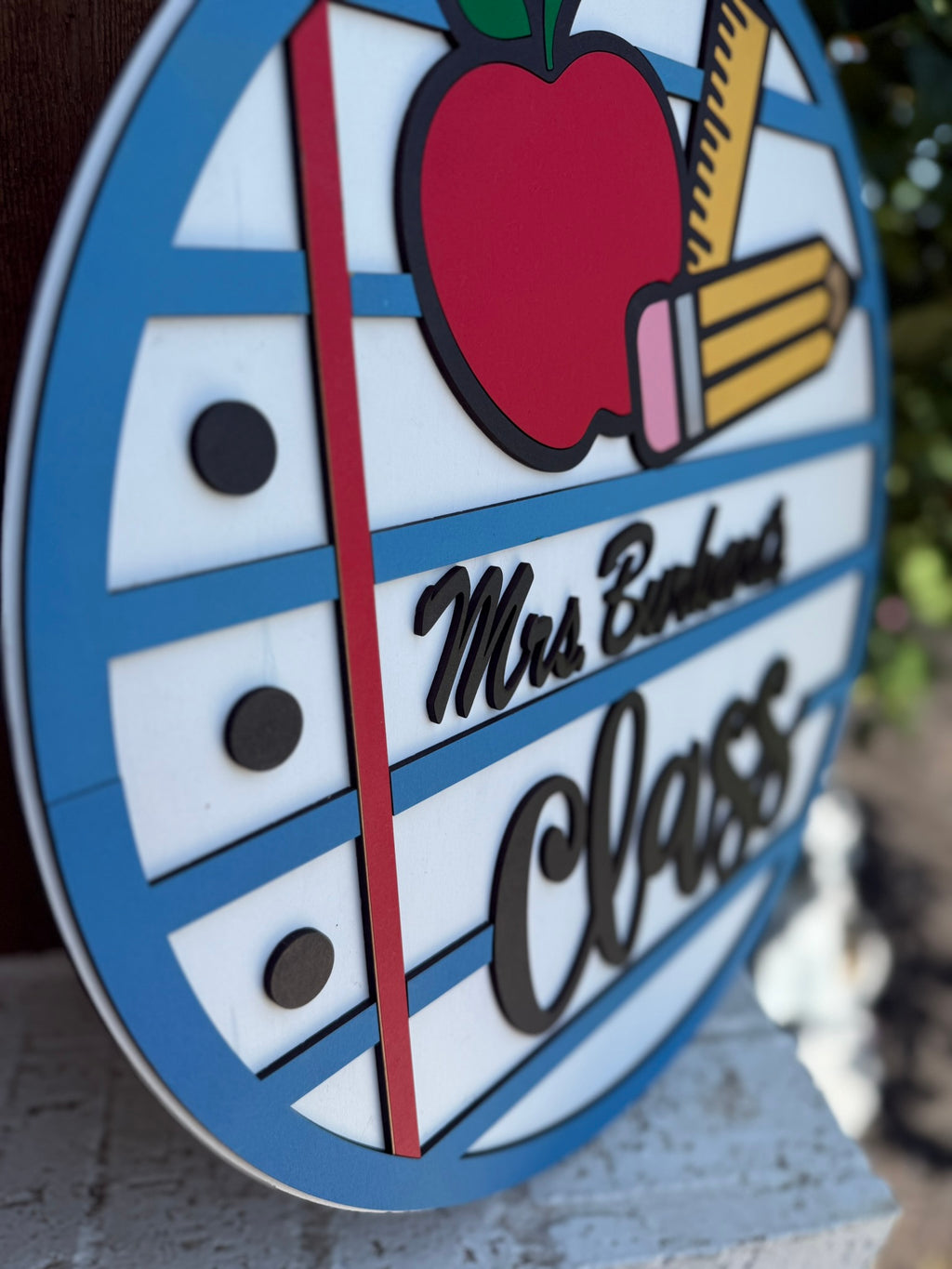 A circular sign resembling lined notebook paper with an apple, two pencils, and the words “Mrs. Barlow’s Class” in script, attached to an outdoor surface.