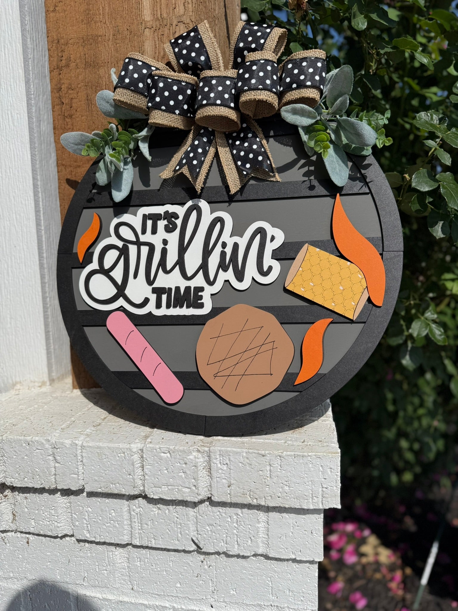 A round sign decorated with black and white bows, greenery, and cutouts of grilled food with the phrase “It’s Grillin’ Time” in white on a dark background, hanging on a brick wall outdoors.