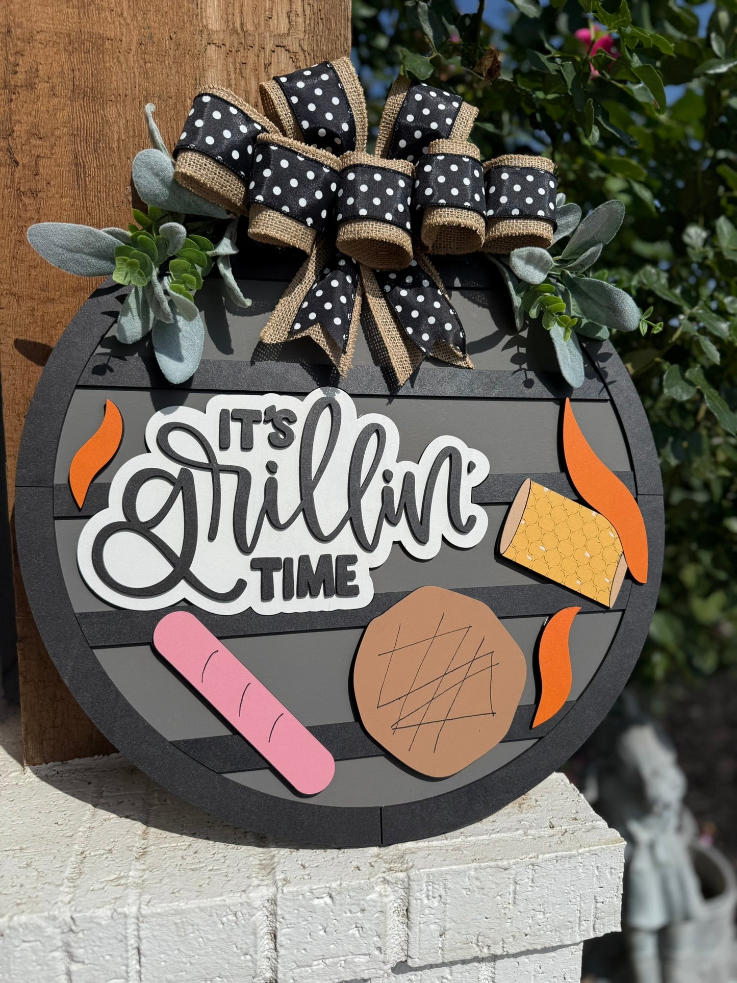 The Its Grillin Time Door Sign Door Wreath is a round sign with black stripes, white letters, a polka-dot bow, faux greenery, and cutouts of steak, sausage, corn, and flames. Shown outdoors leaning against a post.