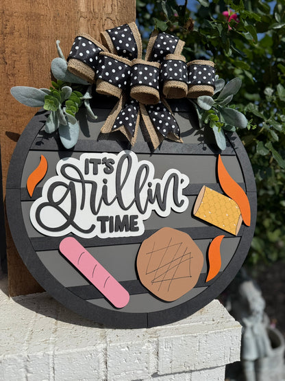 The Its Grillin Time Door Sign Door Wreath is a round sign with black stripes, white letters, a polka-dot bow, faux greenery, and cutouts of steak, sausage, corn, and flames. Shown outdoors leaning against a post.